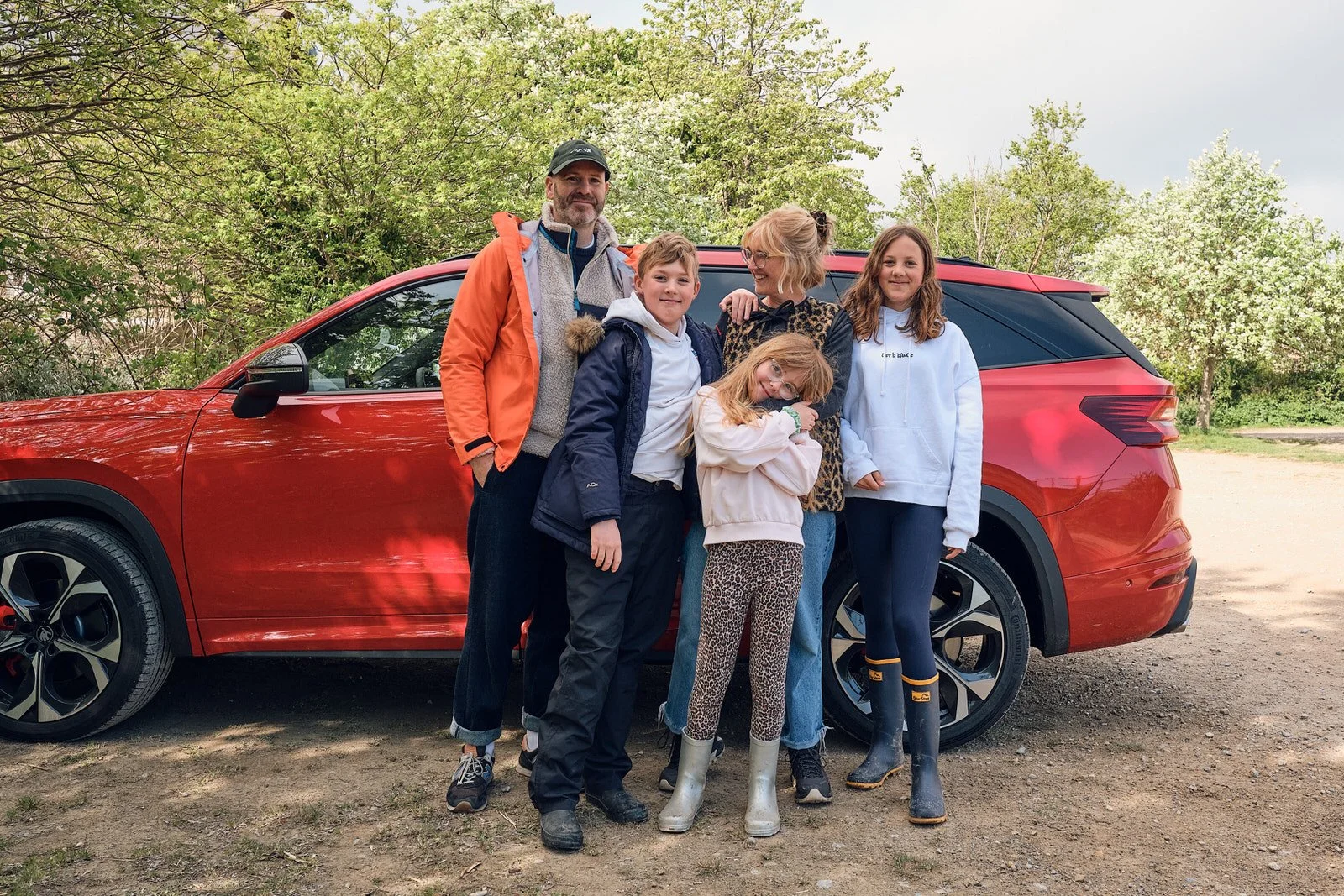 A family of six standing outdoors in front of a red SUV, with trees and a cloudy sky behind them. The family includes two adult men and four children, all smiling and dressed in casual clothing, some wearing jackets and rain boots.