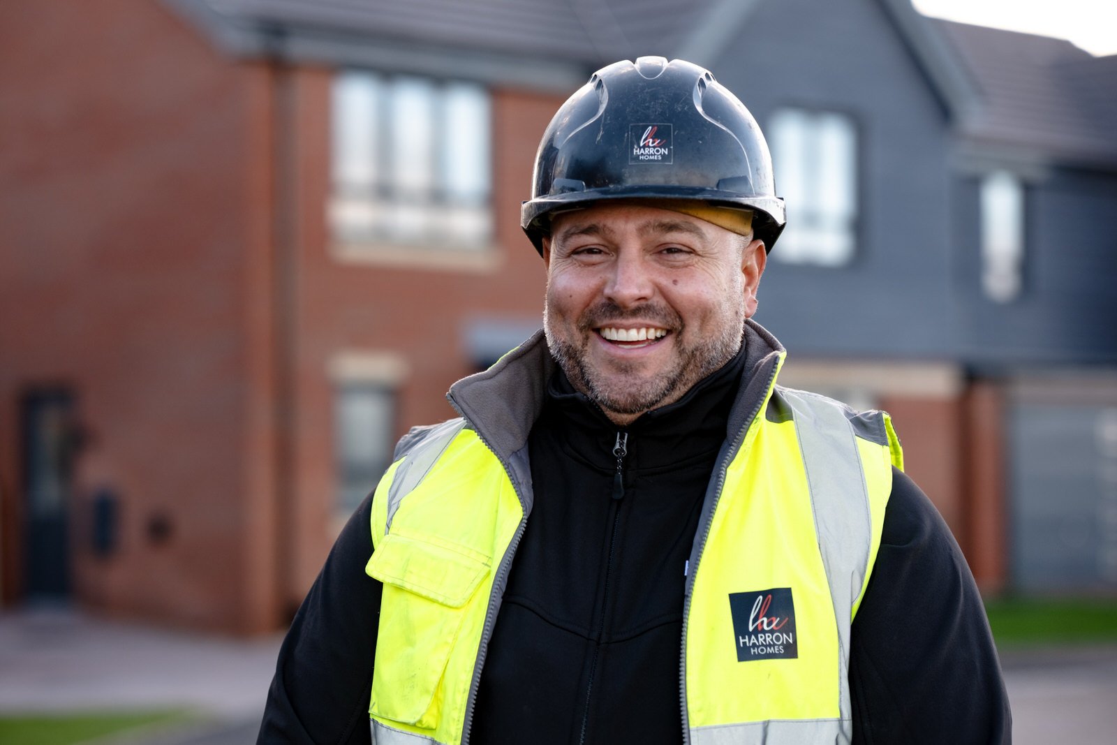 A construction worker wearing a black hard hat and a yellow safety vest with the Harron Homes logo, standing outside in front of residential buildings, smiling.