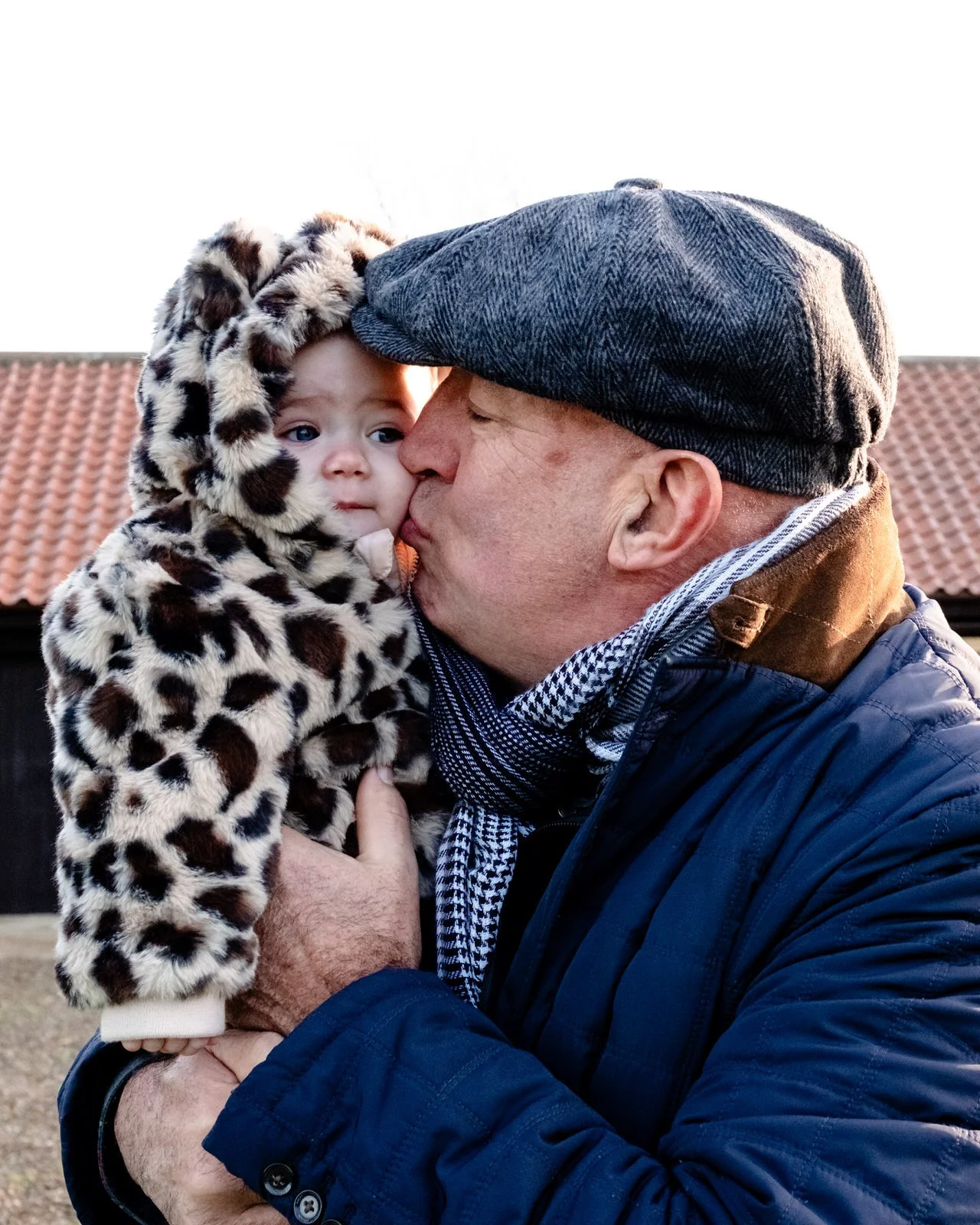 A man in a blue jacket and gray flat cap kisses a young child in a leopard print coat with a hood, outdoors in front of a building with a tiled roof.