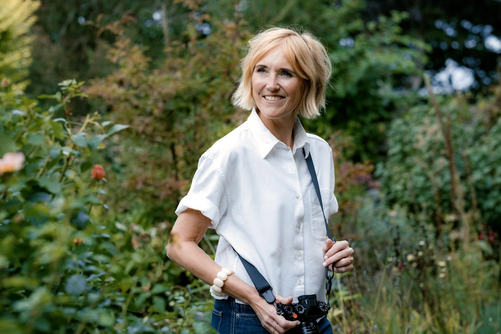 A woman with blonde hair smiling and holding a camera in a green outdoor setting with trees and plants.