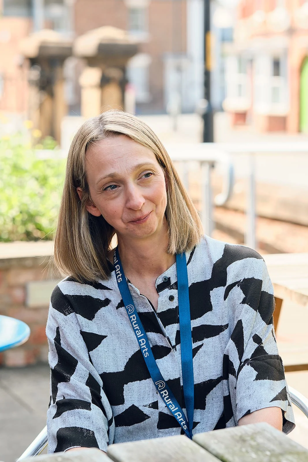 A woman with shoulder-length blonde hair wearing a black and white patterned shirt and a lanyard with 'Rural Arts' written on it, sitting outdoors with a blurred background of buildings and greenery.