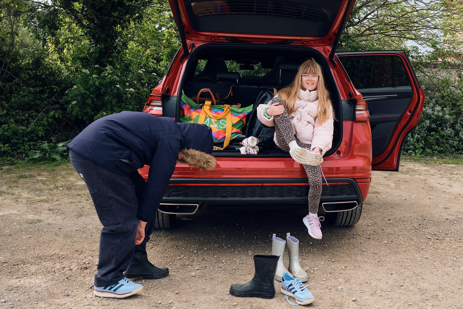 A family of six standing outdoors in front of a red SUV, with trees and a cloudy sky behind them. The family includes two adult men and four children, all smiling and dressed in casual clothing, some wearing jackets and rain boots.