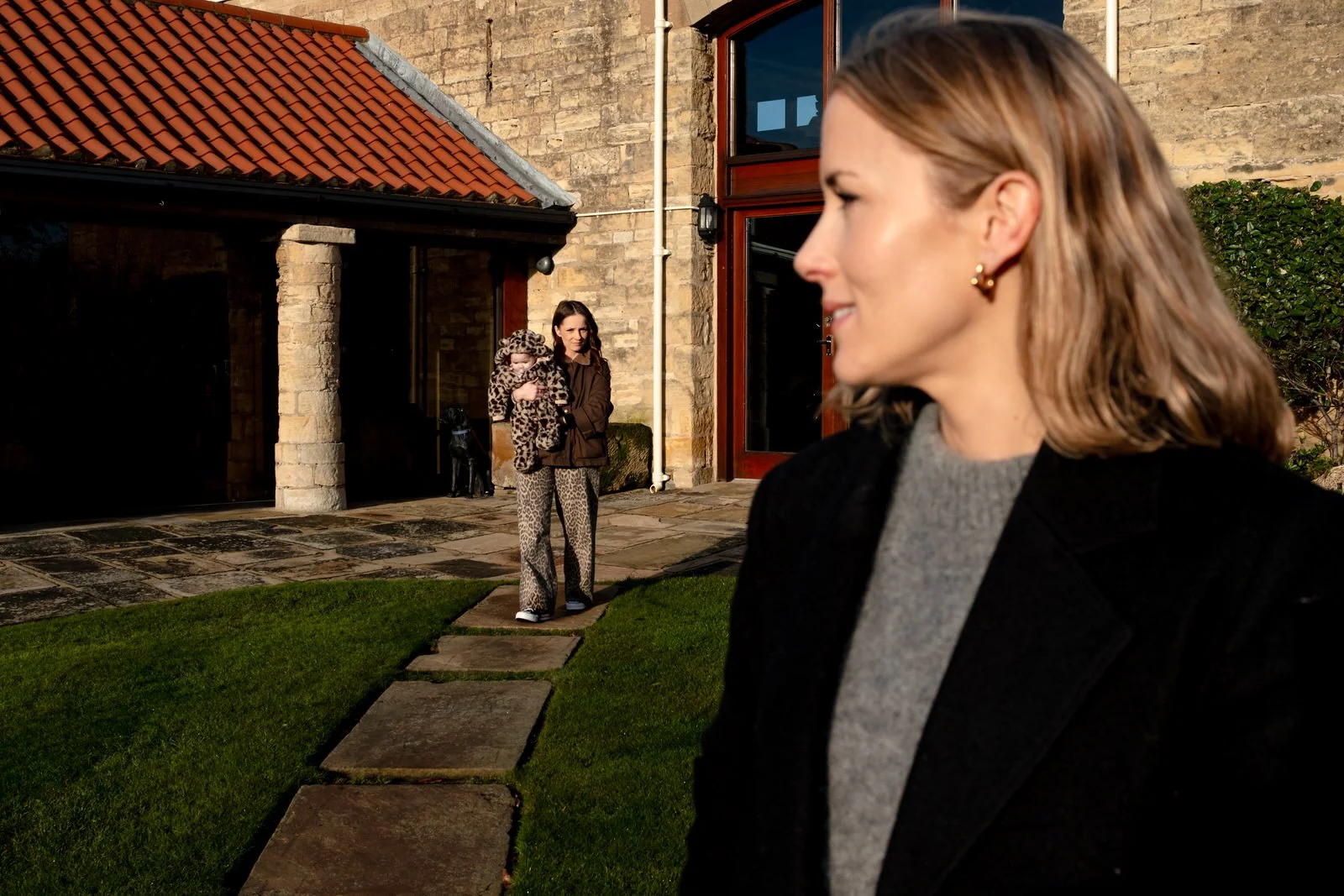 A woman in the foreground is smiling and looking to her right, wearing a black jacket and a gray sweater. In the background, another woman is holding a child dressed in a leopard-print outfit, standing on a stone pathway outside a brick house with a 
