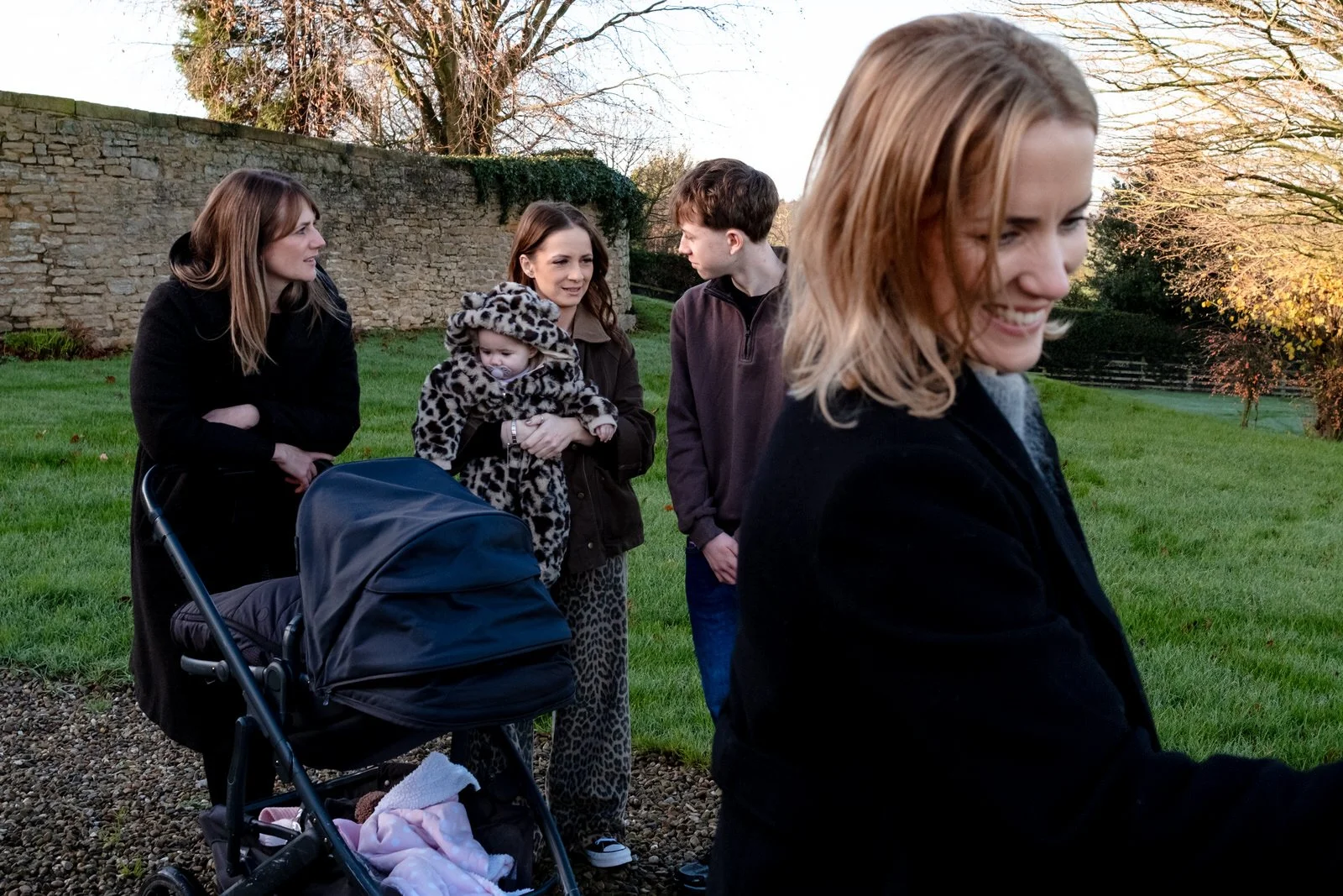 A group of five people, including two young women, a man, and two children, standing outdoors on a grassy area with a stone wall and trees in the background. One woman is pushing a stroller, and one girl is holding a baby in a leopard-print coat. The