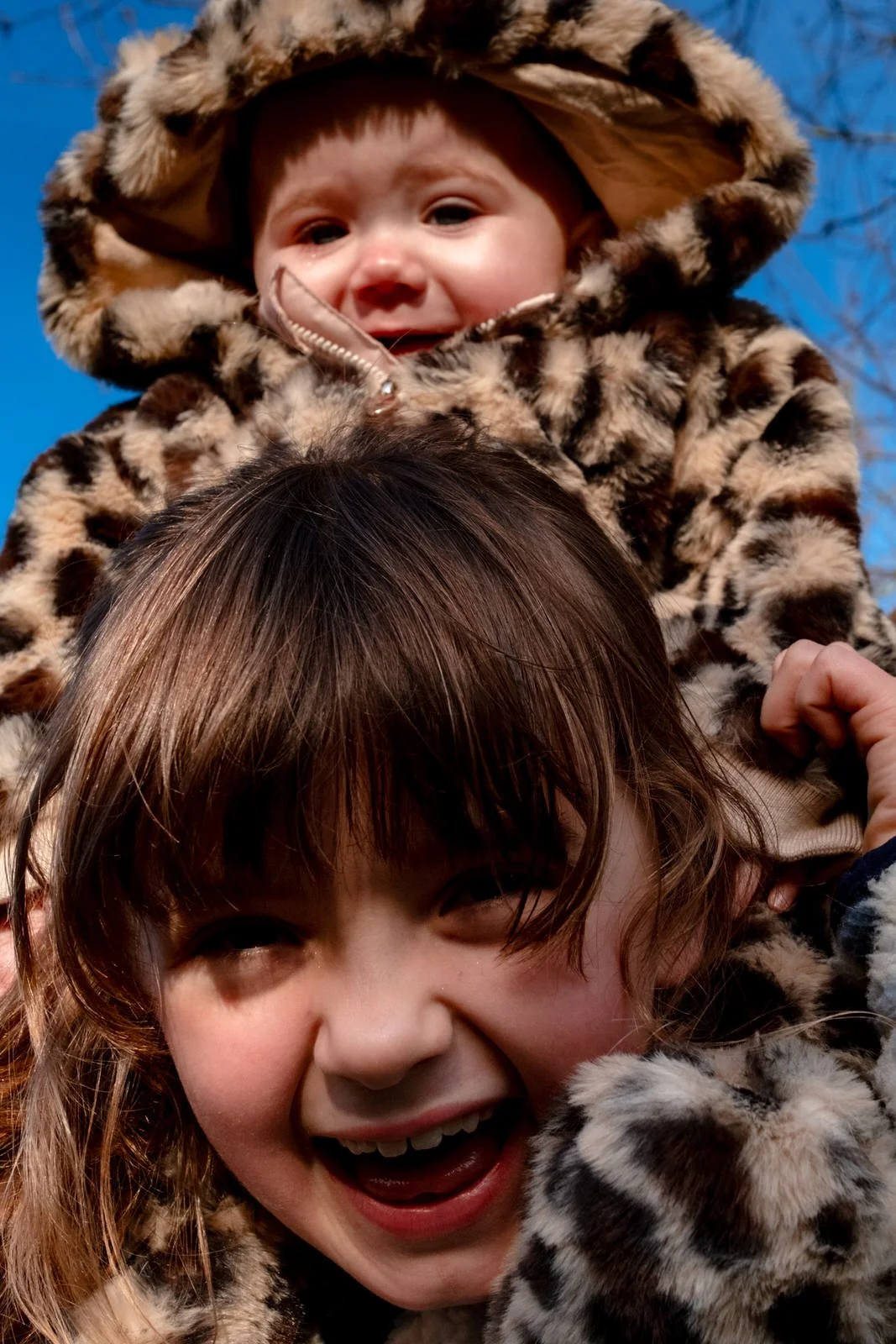 Two children, a girl and a boy, smiling and playing outside on a clear day, with the boy sitting on the girl's shoulders. They are dressed in matching leopard print jackets.
