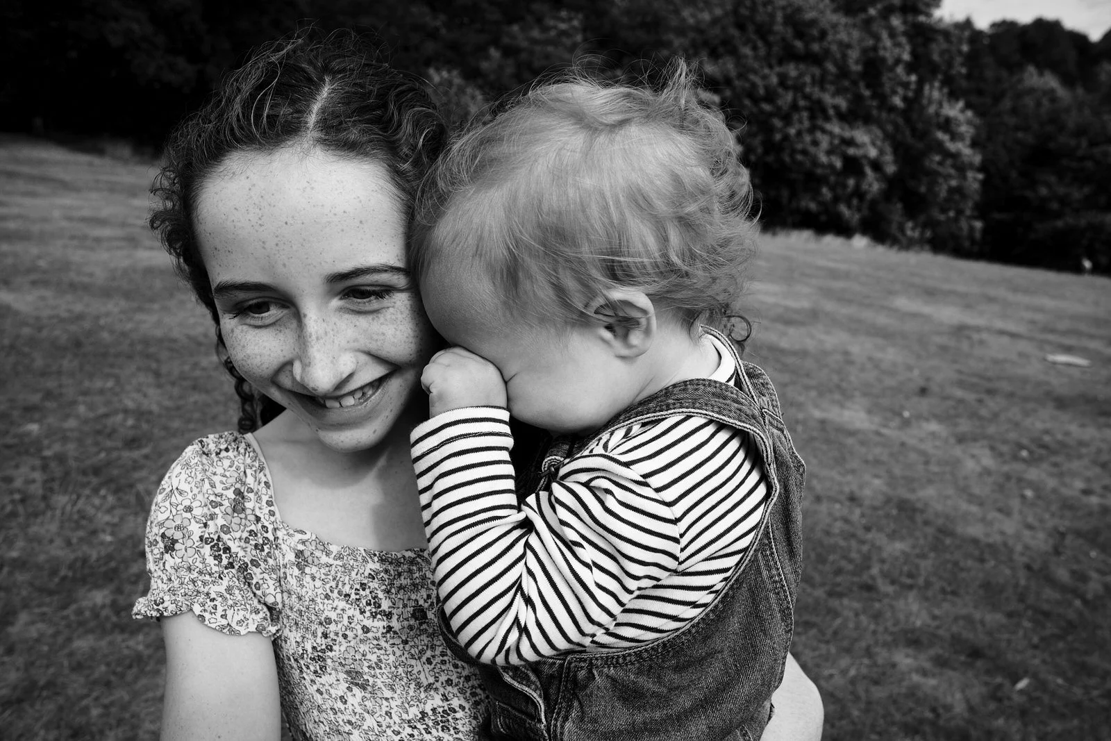 A young girl holding a small child in an outdoor setting with trees in the background.