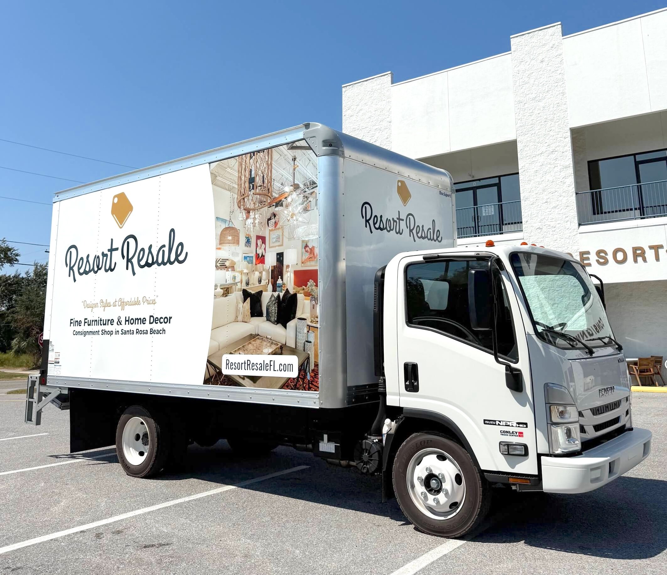 A white truck with an advertisement on its side for Resort Resale, featuring furniture and home decor, parked in a lot outside a white building with balconies and the word 'RESORT' partially visible.