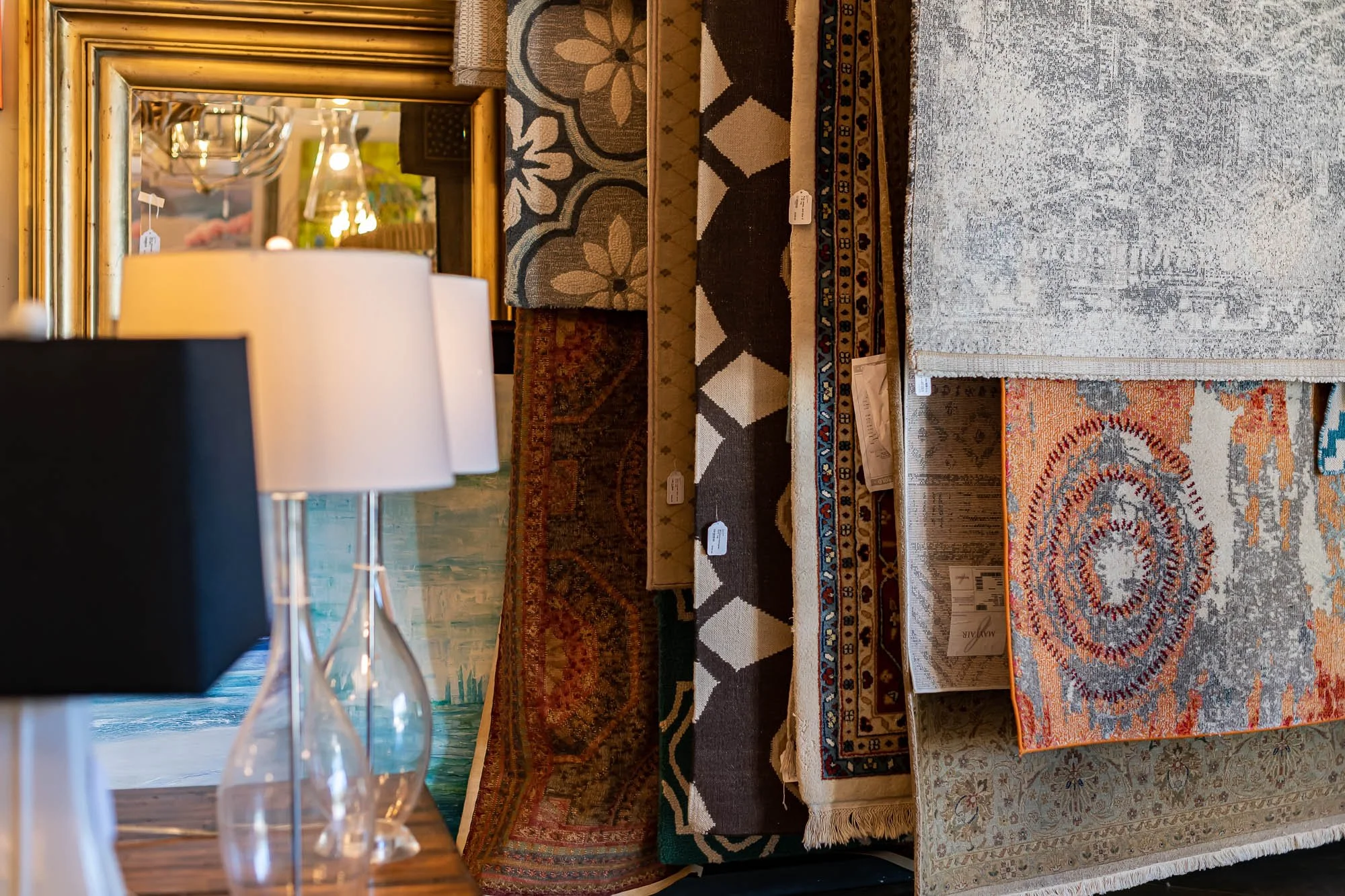Stacked area rugs in various colors and patterns displayed in a home decor store, with a table lamp with a white shade and clear glass base in the foreground.