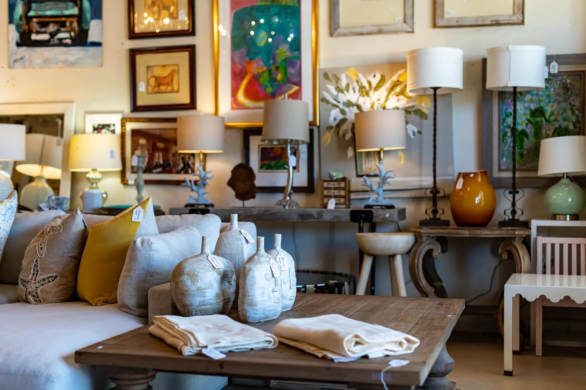 A gallery of various framed paintings and artwork on a wall, surrounded by table lamps and decorative objects in a room filled with furniture and decor.