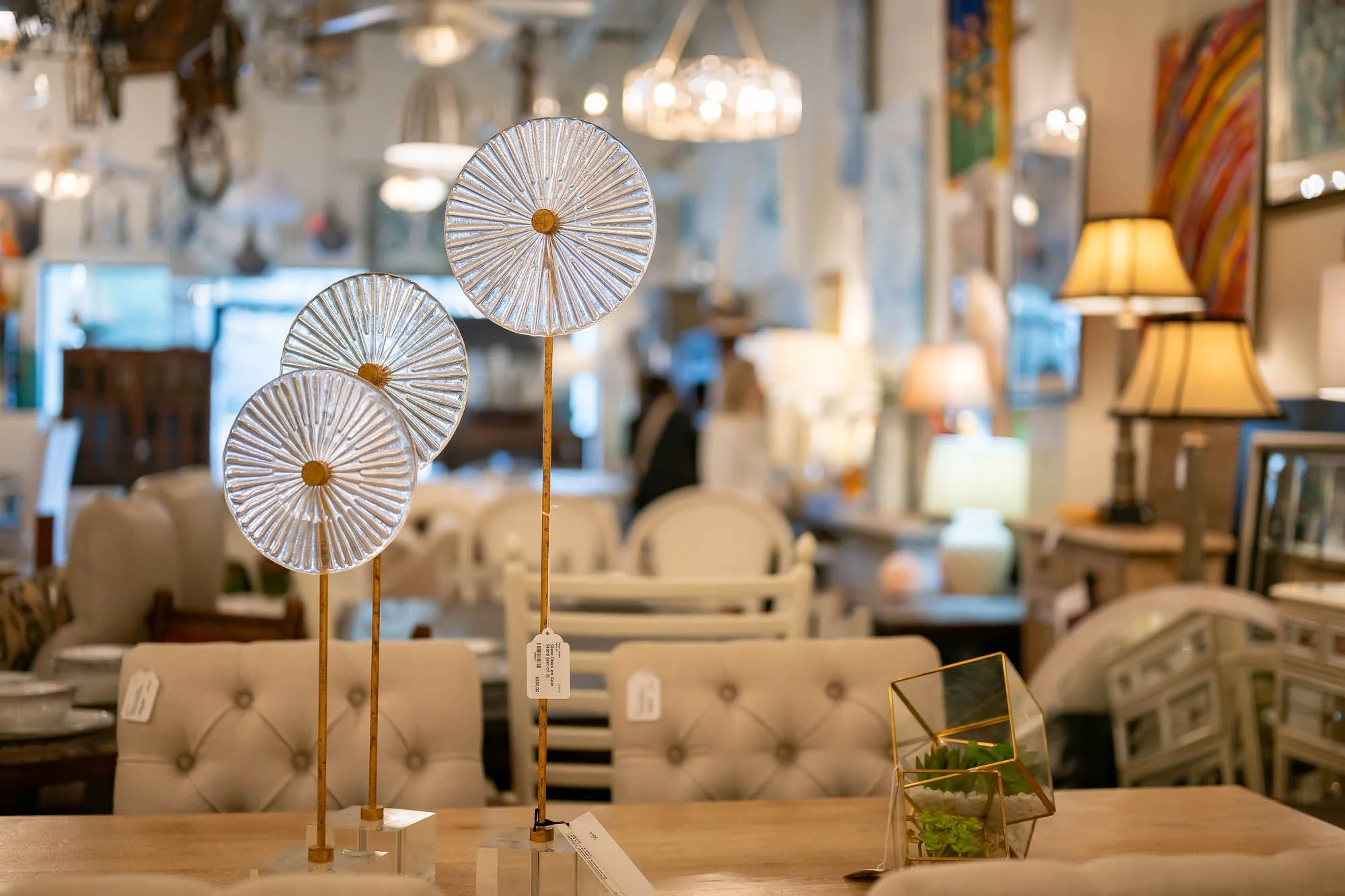 Decorative glass and metal sculptures of three circular, sunburst-style objects on tall sticks on a wooden table. The background shows a furniture store interior with various lamps, chairs, and artwork.