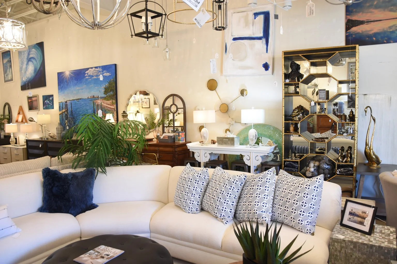 Interior of a furniture store showcasing a white sectional sofa with patterned pillows, surrounded by various lamps, artwork, shelves with decorative items, and a large green plant.