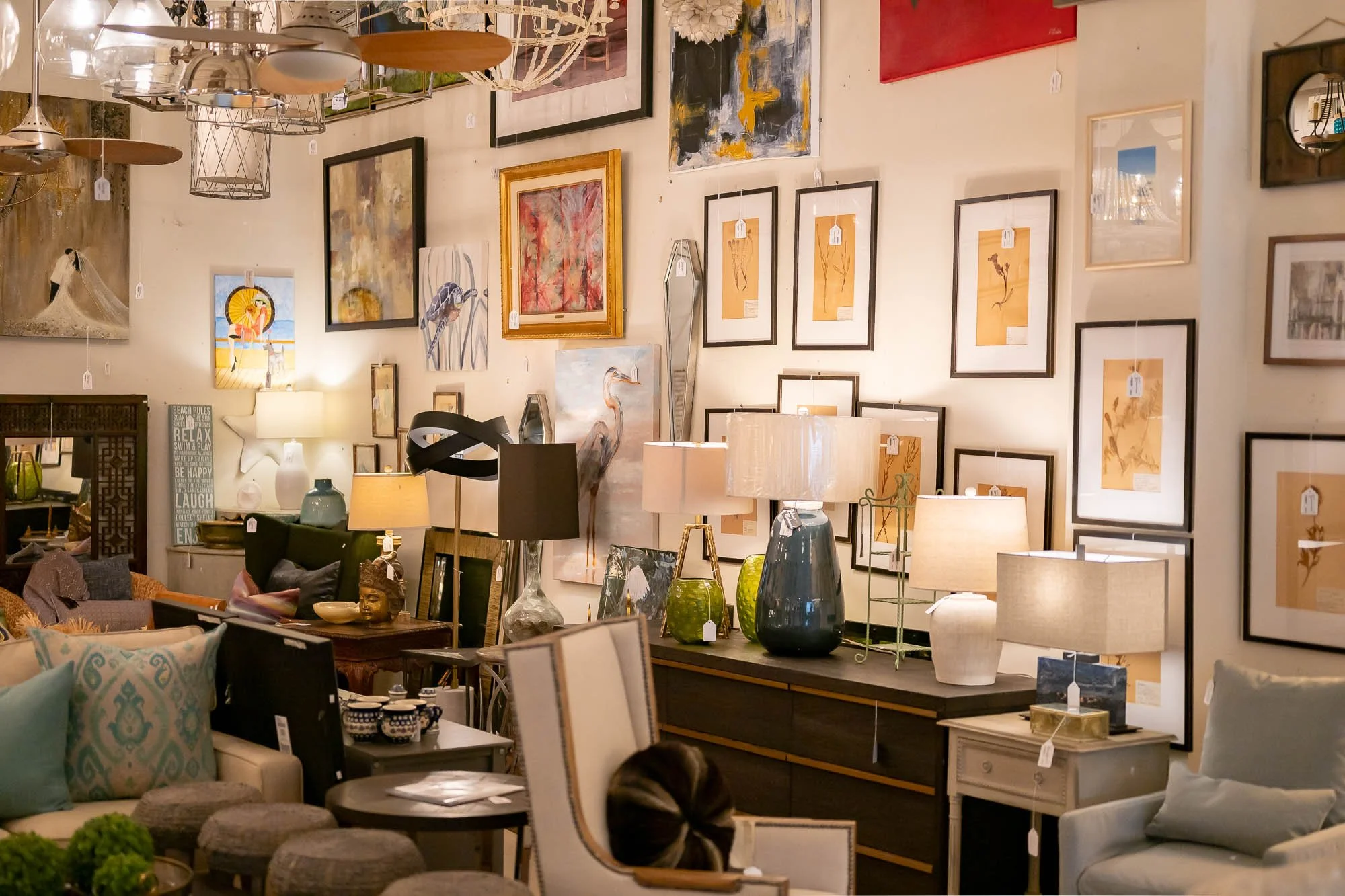 Interior of an art gallery or furniture store displaying various paintings and framed artwork on the wall, along with lamps and decorative items on furniture pieces.