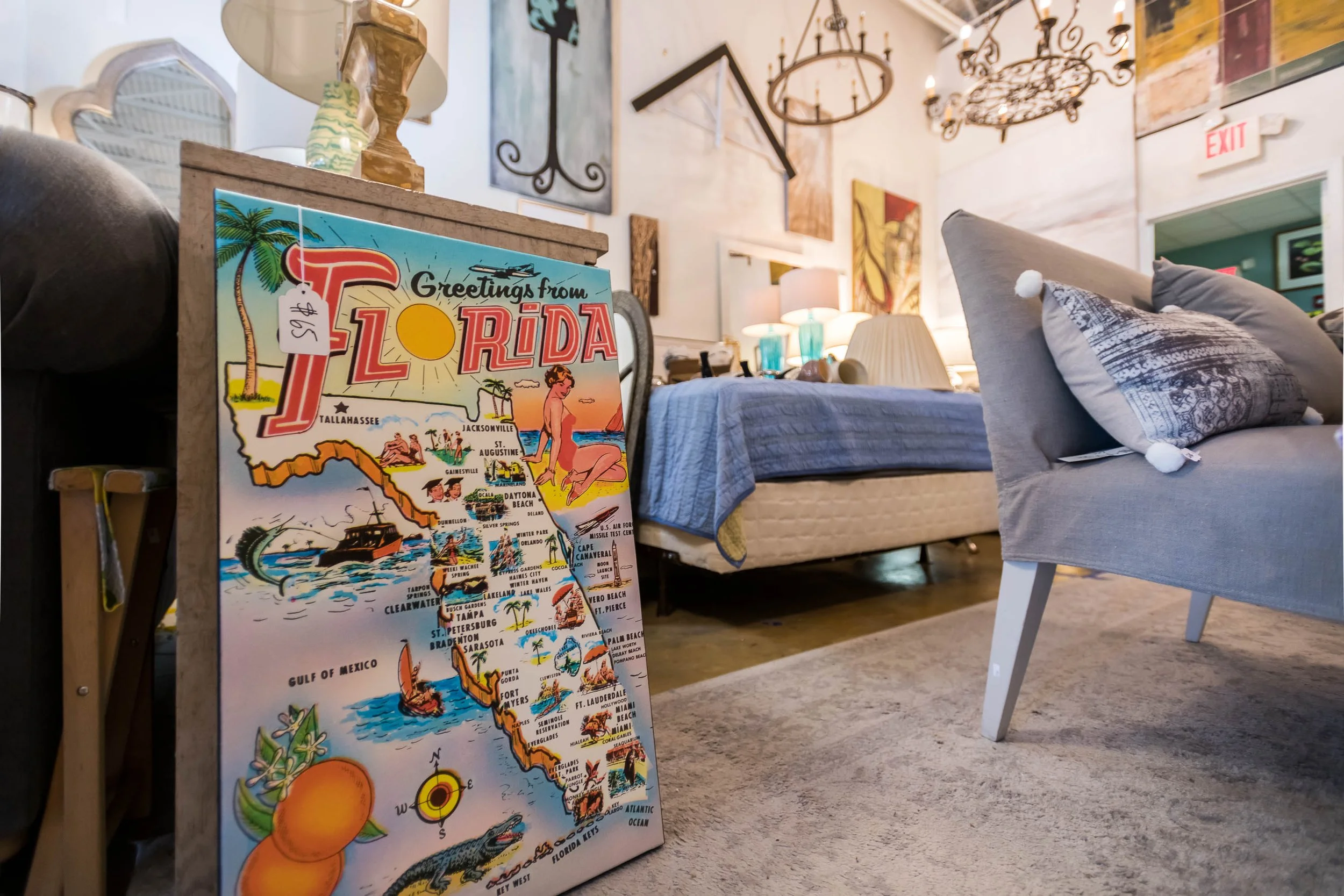A colorful vintage postcard of Florida leaning against a piece of furniture, with a bed, gray cushions, lamps, wall art, and chandeliers in a cozy room.