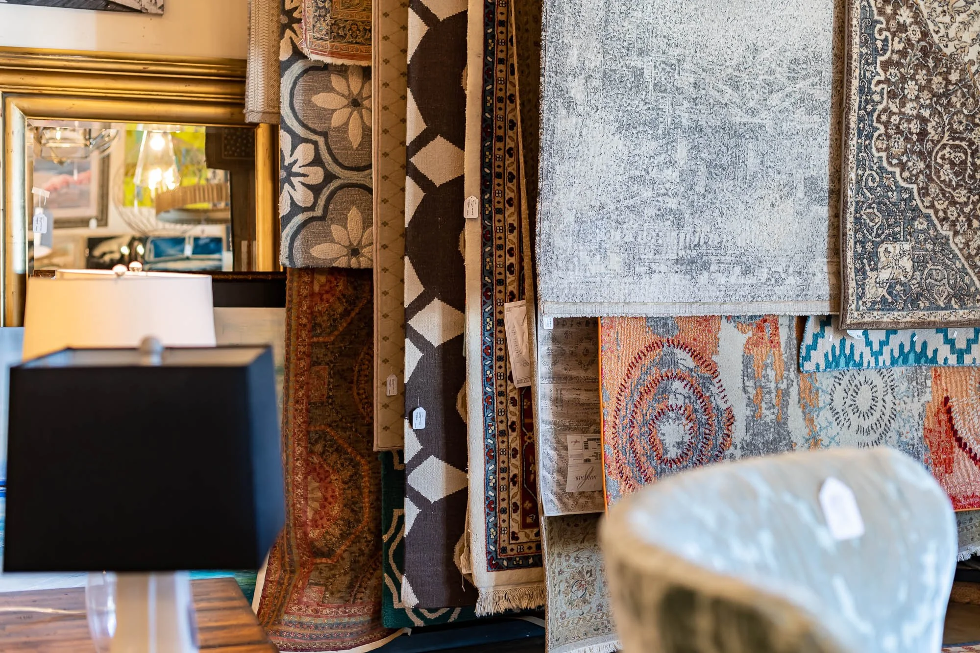 A variety of colorful area rugs layered on top of each other in a room, with a mirror reflecting lamps and furniture, and a black table lamp in the foreground.