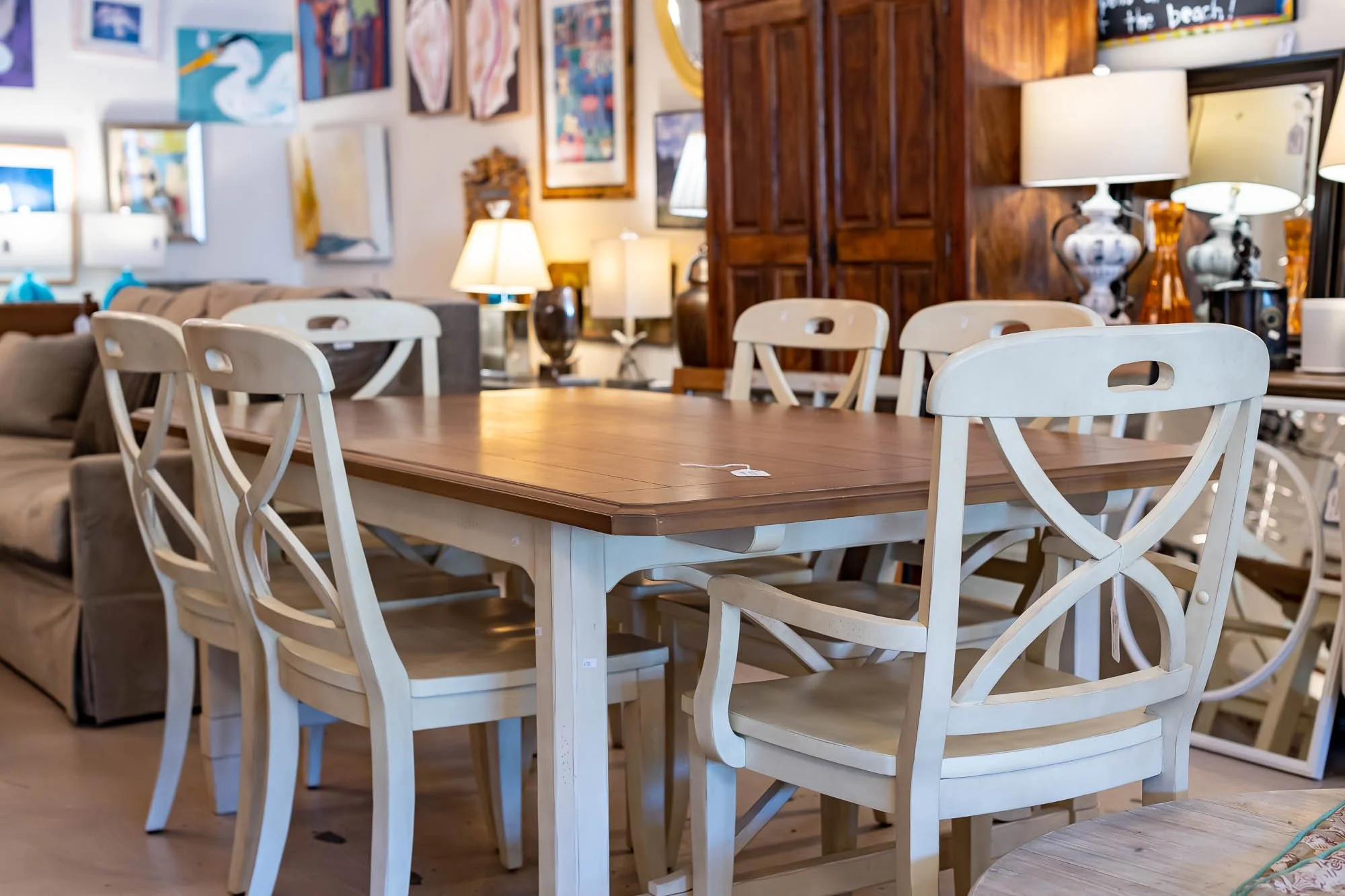 A wooden dining table surrounded by six light-colored chairs in a furniture store, with lamps, artwork, and mirrors on the walls in the background.