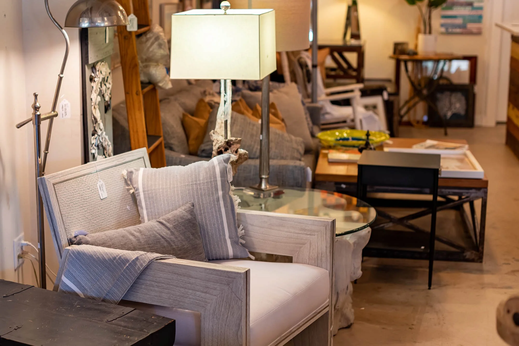 A furniture store displays various living room pieces including a wooden armchair with cushion pillows, a glass top coffee table, a sofa with brown pillows, a standing lamp with a shade, and other chairs in the background.