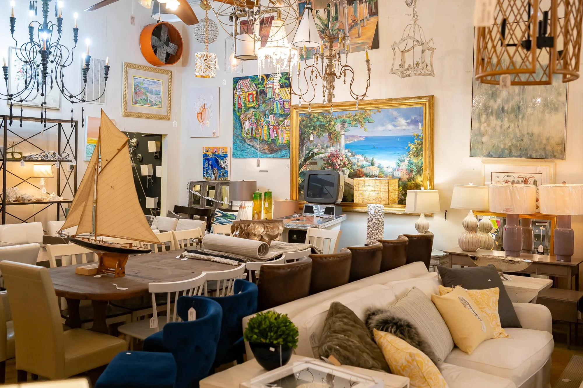 An interior view of a furniture and home decor store showing various paintings, lamps, and decorative items arranged on tables, shelves, and walls.