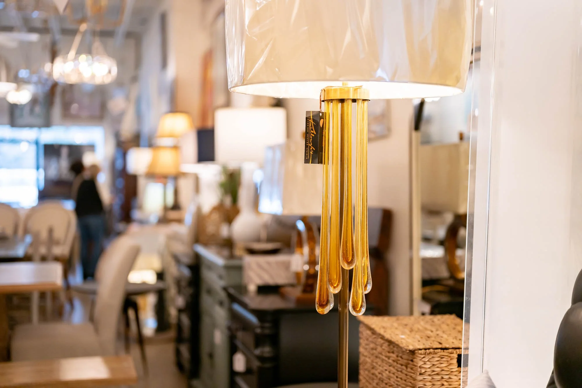 A decorative table lamp with a gold base and glass teardrop accents, positioned in a furniture store with other lamps and home decor items in the background.