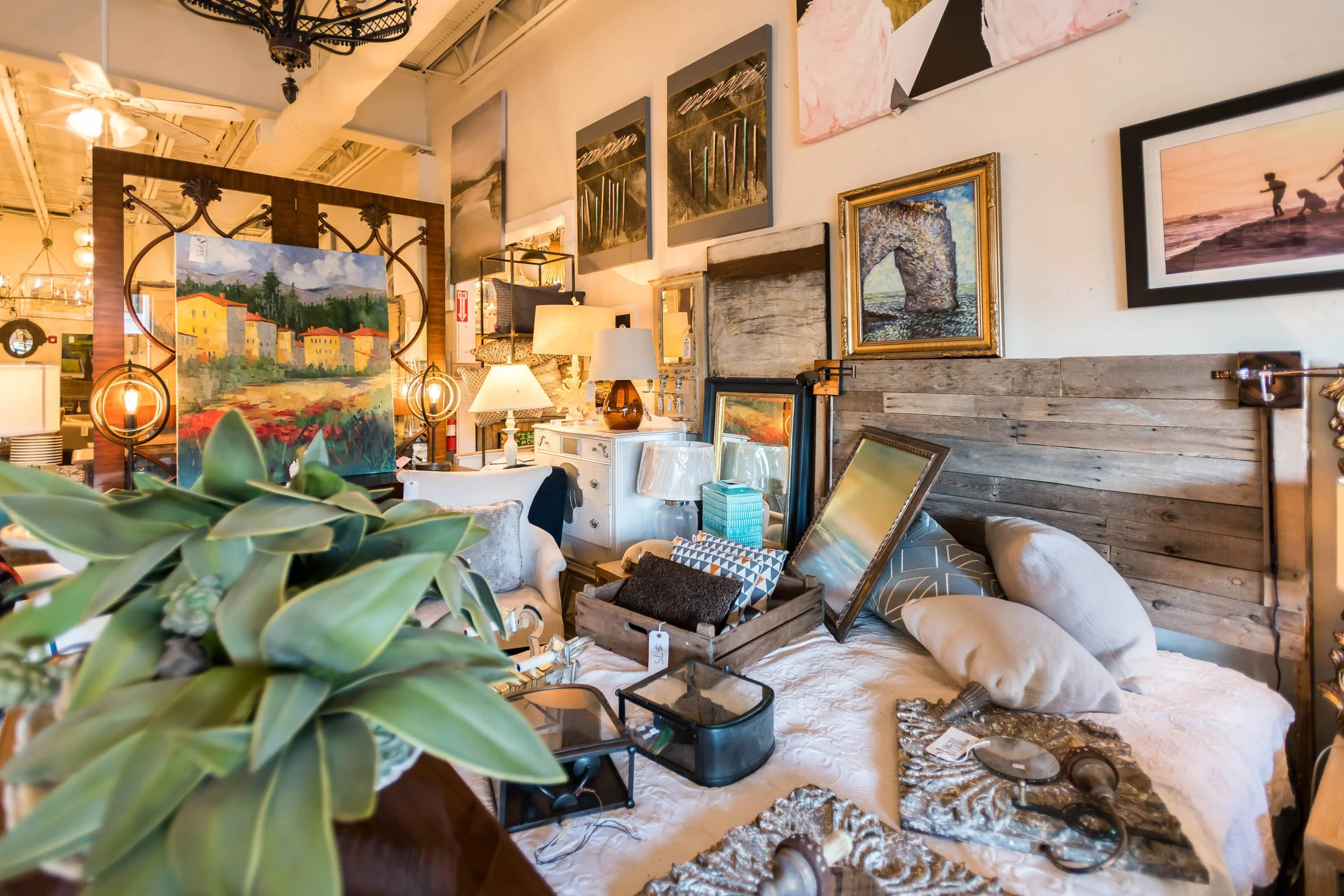 A cozy furniture display in an art gallery or antique shop, featuring a bed, various paintings, lamps, mirrors, and decorative items.