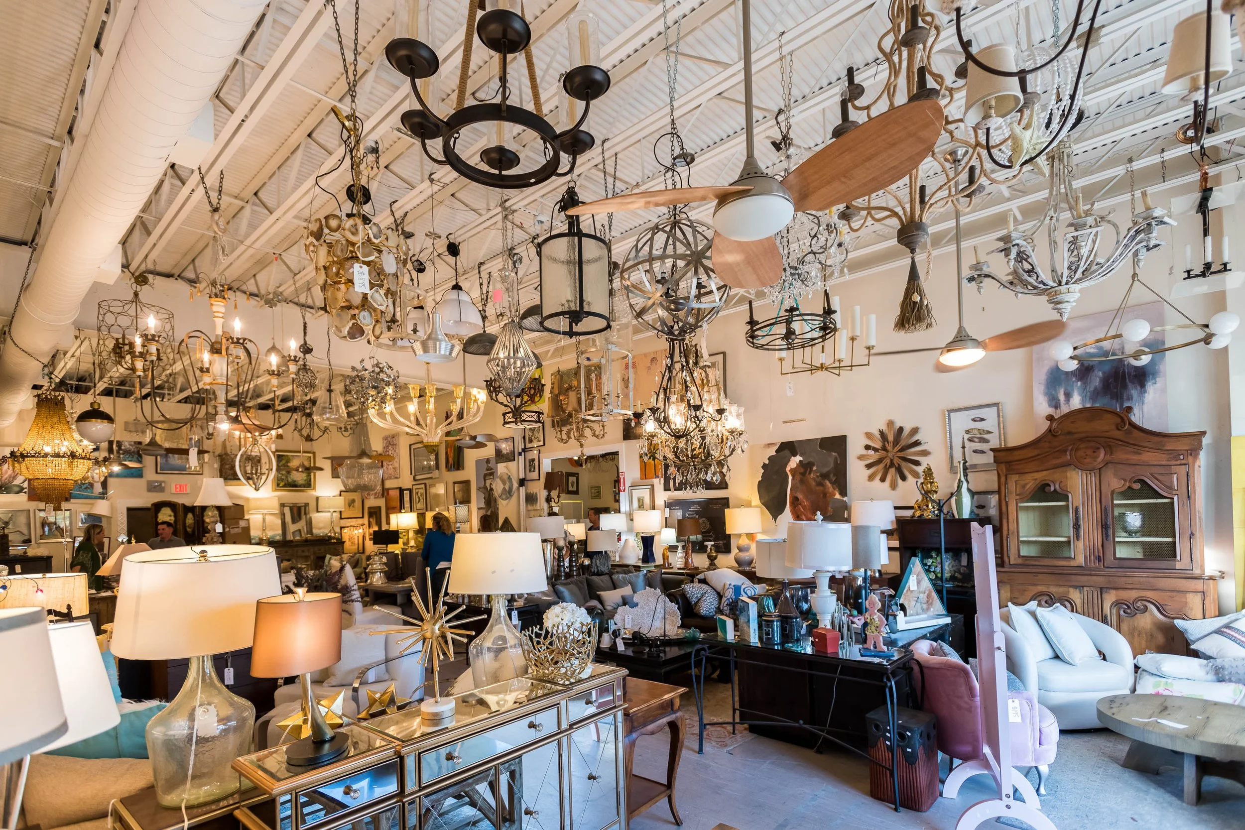 Interior of a vintage furniture and lighting store with various chandeliers and lamps hanging from the ceiling, and furniture and decor items displayed throughout the space.