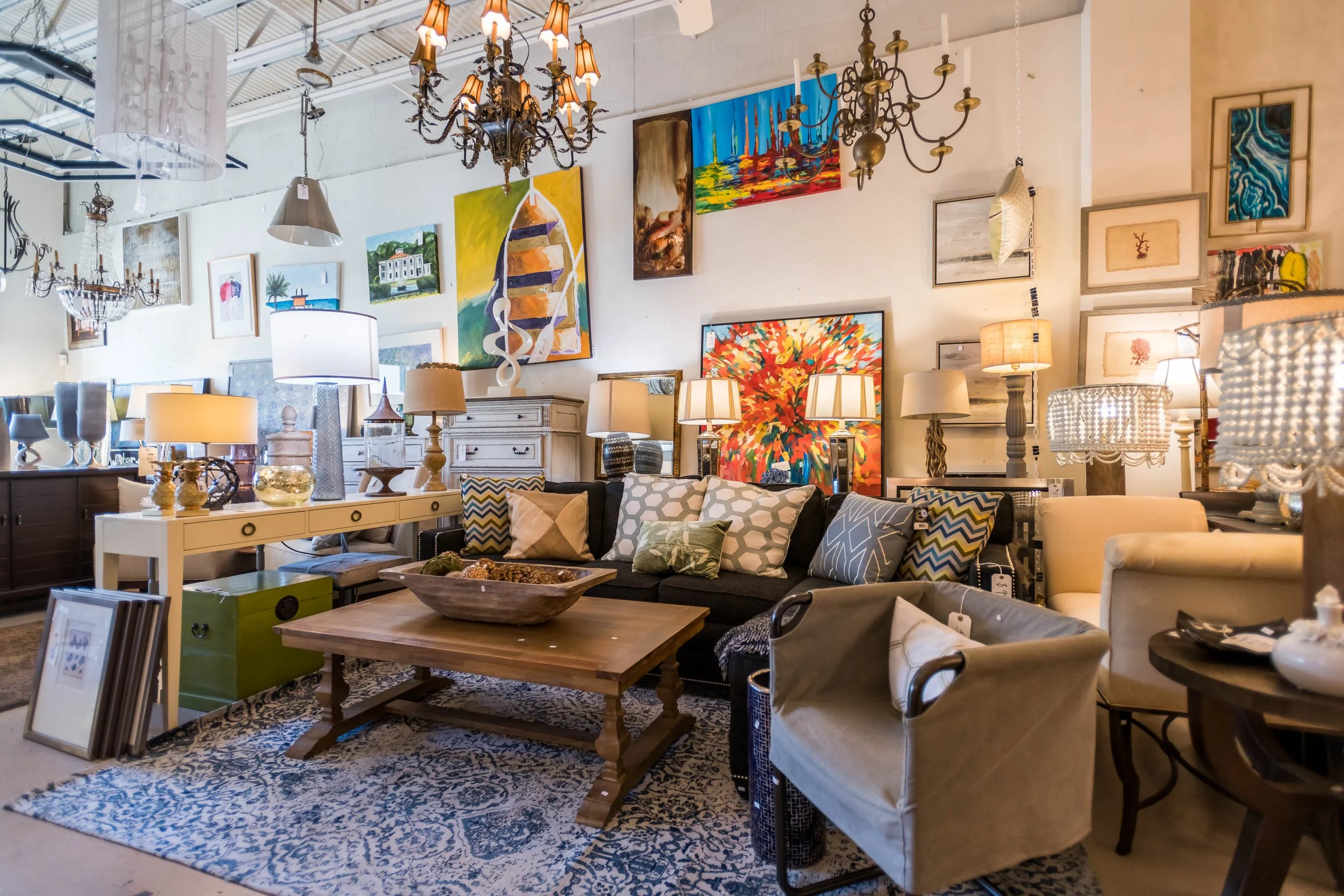 A furniture store display featuring a variety of lamps, art paintings on the wall, a black sofa with patterned pillows, a wooden coffee table, and multiple framed artworks and decorative items, all illuminated by chandeliers and lighting fixtures.