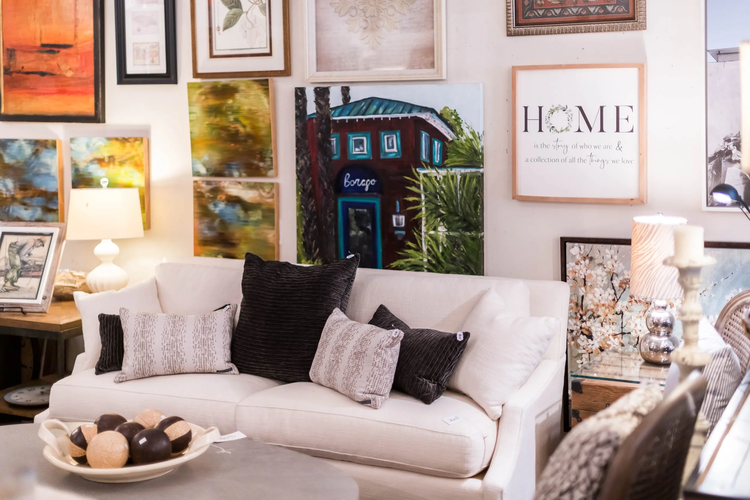 Show room with white sofa, black and patterned throw pillows, surrounded by wall art and paintings, with side tables and lamps.