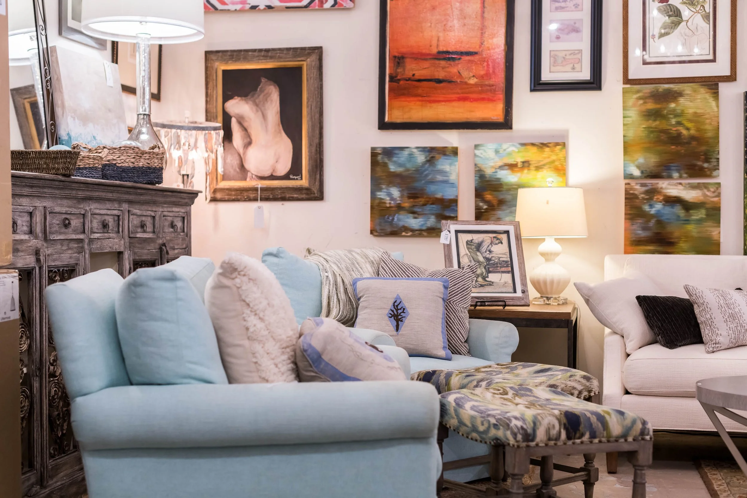 Show room with light blue and white sofas, assorted decorative pillows, artwork on the walls, and a wooden side table with a framed picture and a table lamp.