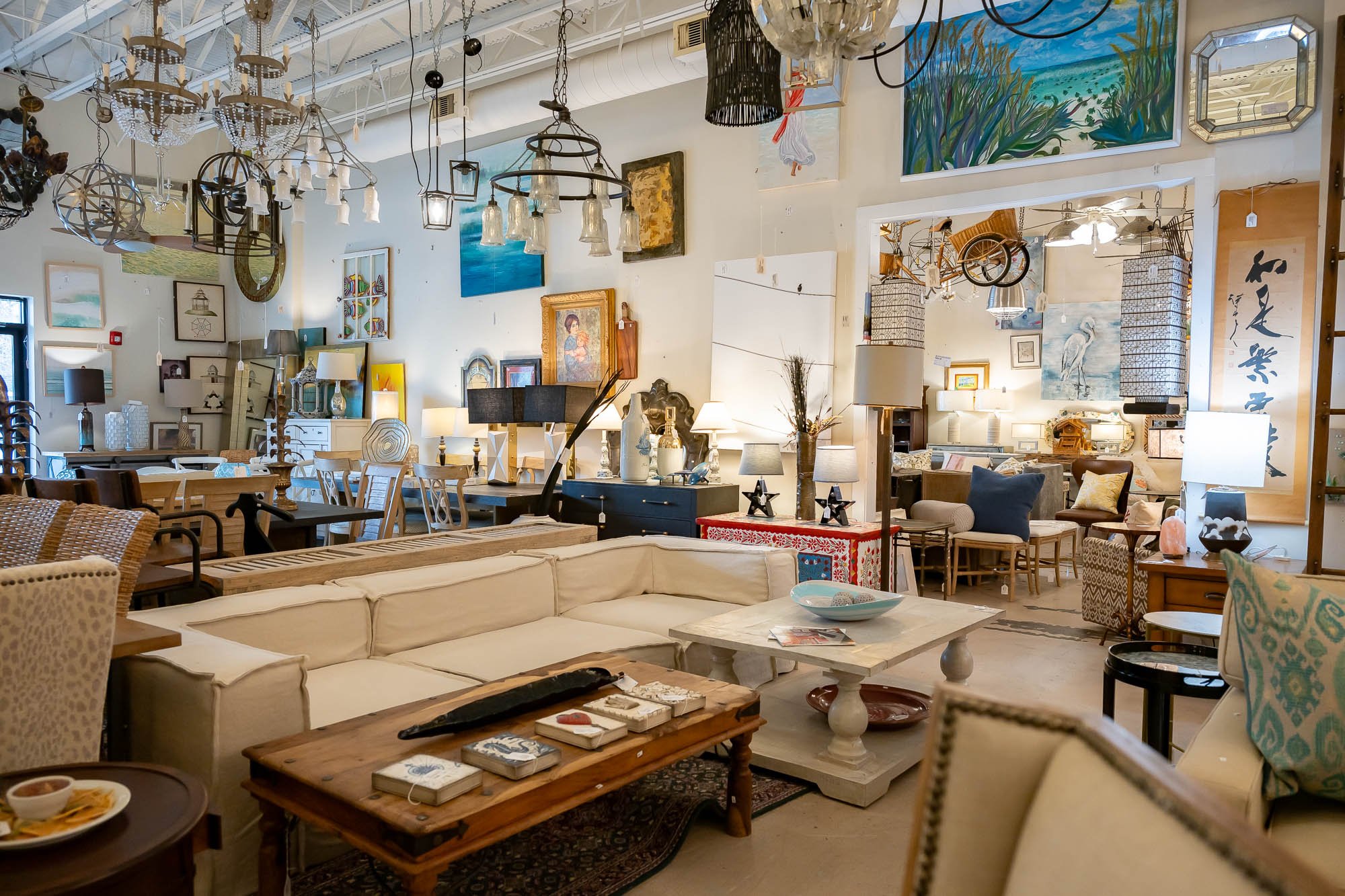 Interior of a furniture store showcasing various sofas, tables, lamps, and decorative art pieces on the walls, with hanging chandeliers and pendant lights.