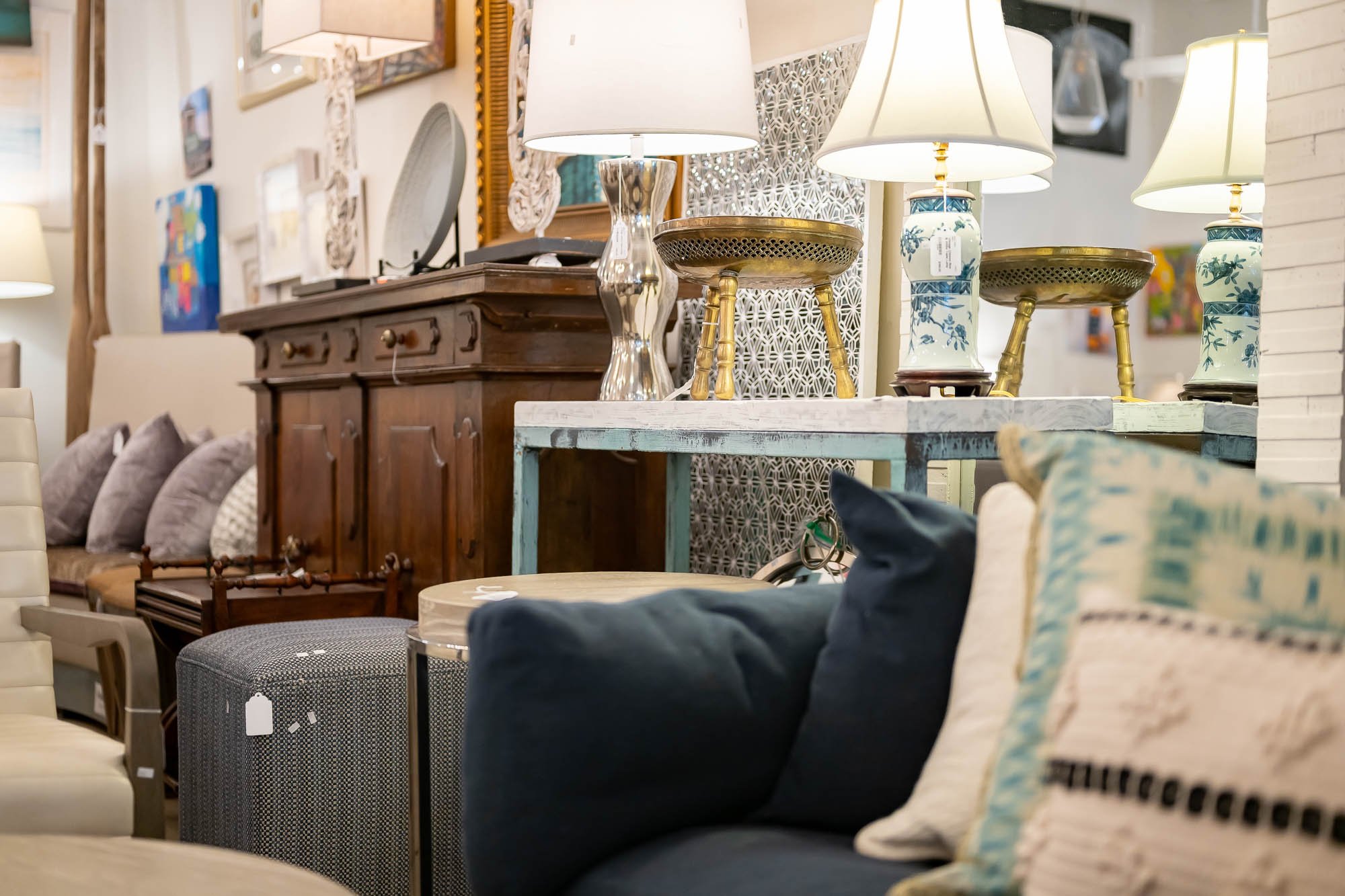 A display of various lamps and furniture in a store, including table lamps with decorative bases, wooden cabinets, and cushioned chairs with pillows.