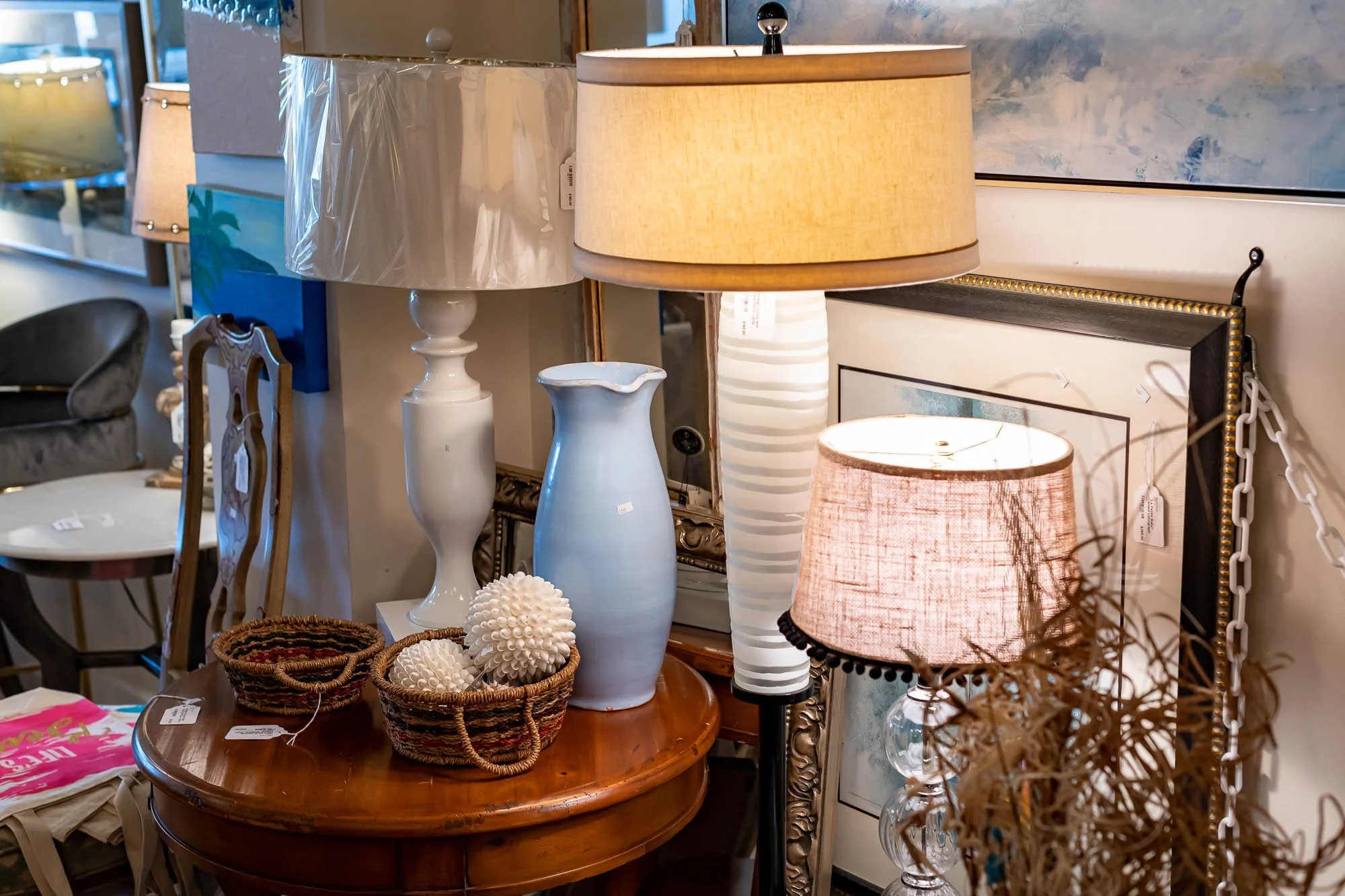 Display of various lamps, vases, and decorative items in a store or gallery setting with framed artwork and furniture in the background.
