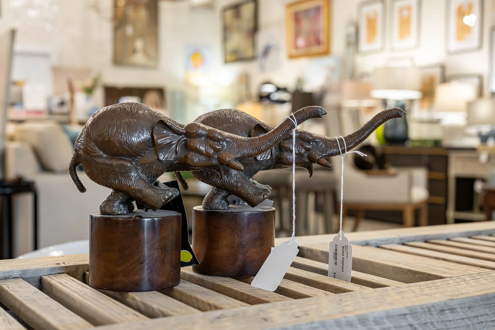 Two elephant sculptures made of dark material on wooden bases, displayed on a wooden pallet in a store interior with framed pictures and furniture in the background.