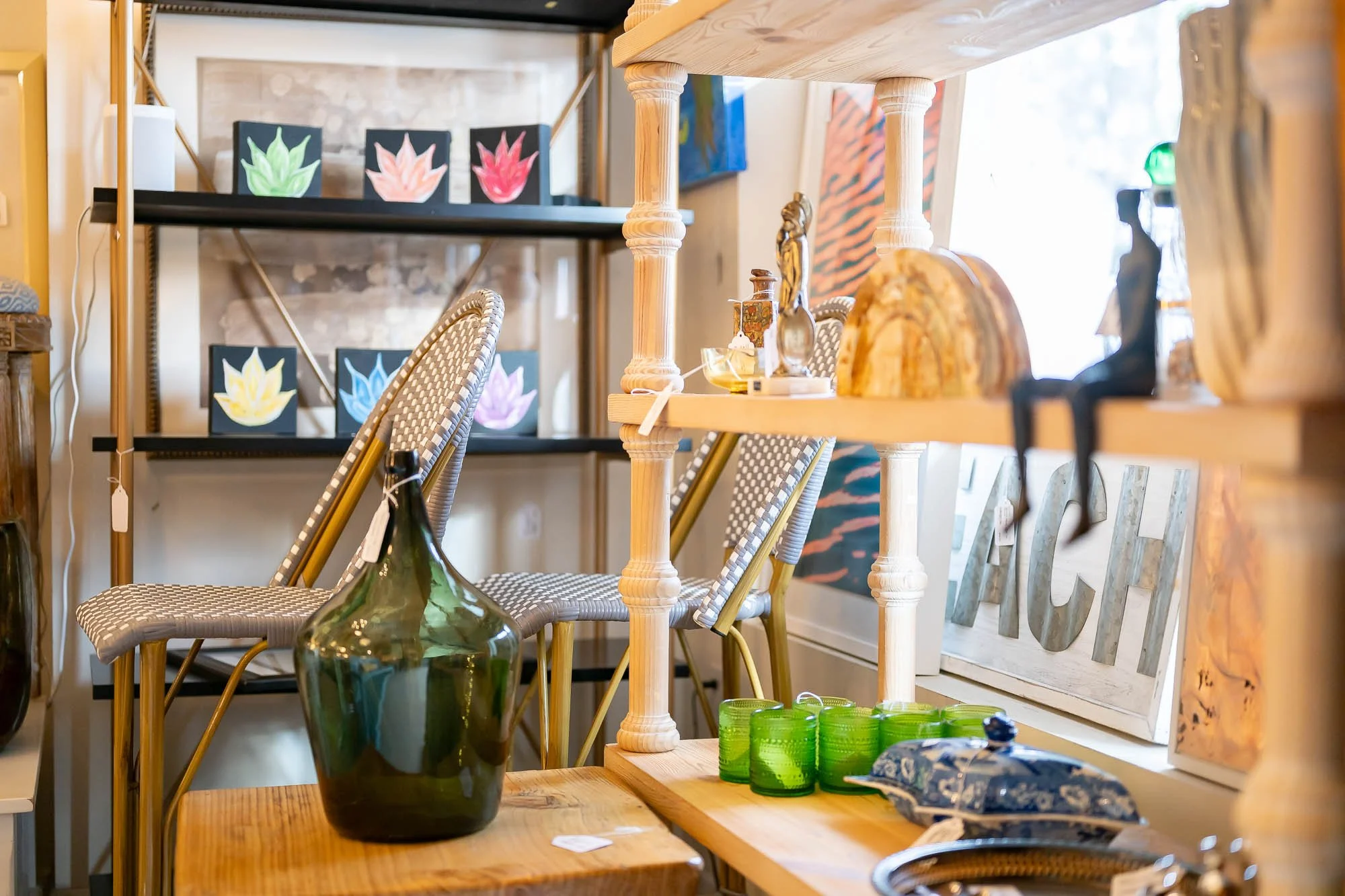 Interior of a store with wooden shelves displaying decorative items, including a large green glass bottle, small green glass cups, figurines, and artwork of colorful lotus flowers on black canvases, with chairs and a sign that says 'BEACH' in the bac