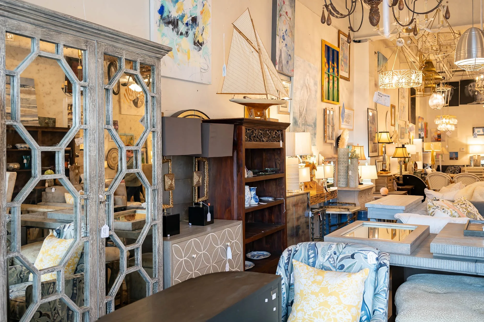 Interior of a furniture store with various lamps, chairs, and decorative items, including a model sailboat on a wooden shelf, and wall art.