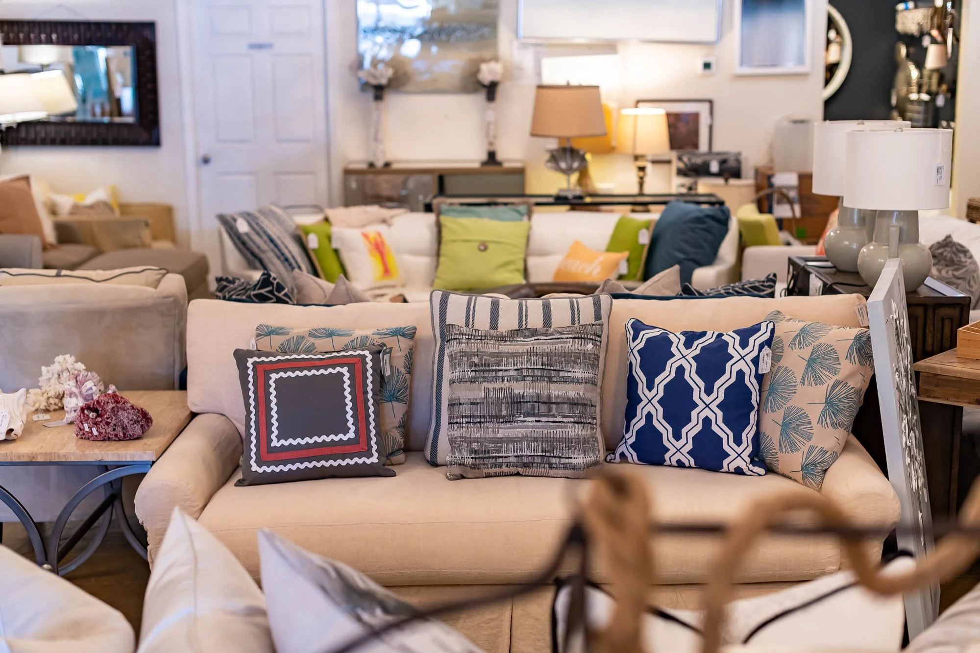 A beige sofa with three decorative pillows featuring various patterns in front of a larger sofa with colorful cushions, set in a furniture store with lamps, framed art, and other furniture visible in the background.