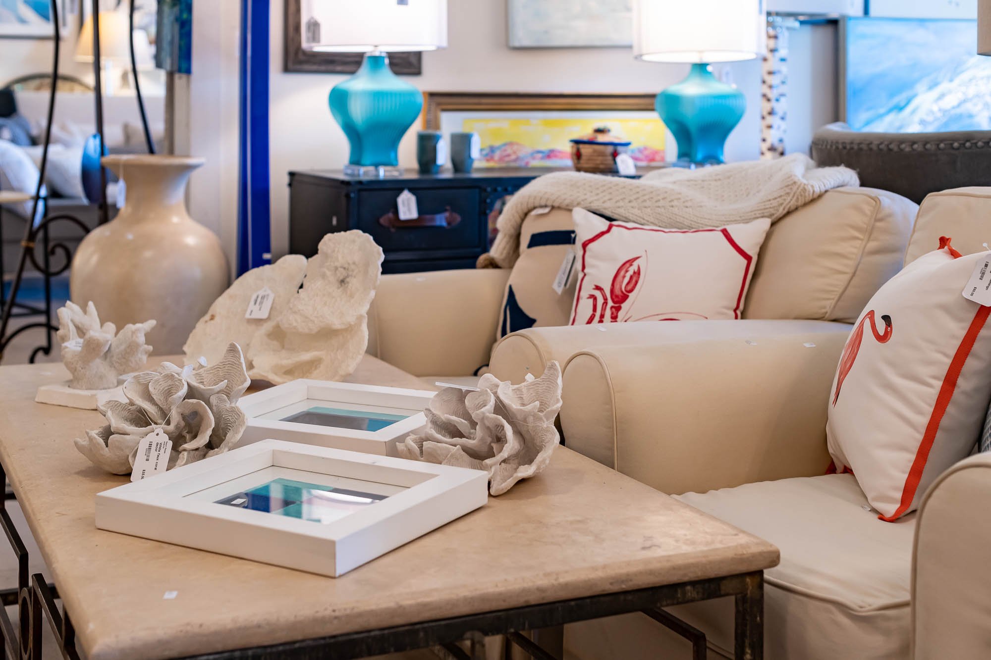 Living room furniture including a beige sofa with lobster-themed pillows and a blanket, a black side table with blue lamps, and a wooden table with coral decor and framed photos, in an interior decor store.
