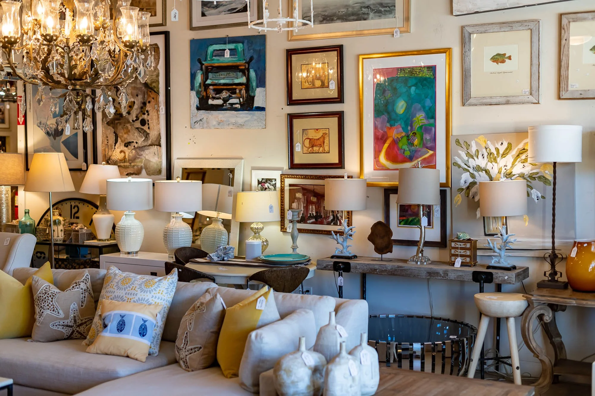 A furniture and decor showroom with a beige sofa decorated with yellow and starfish-print pillows, a variety of table lamps, framed artwork, and decorative vases and sculptures on tables and walls.