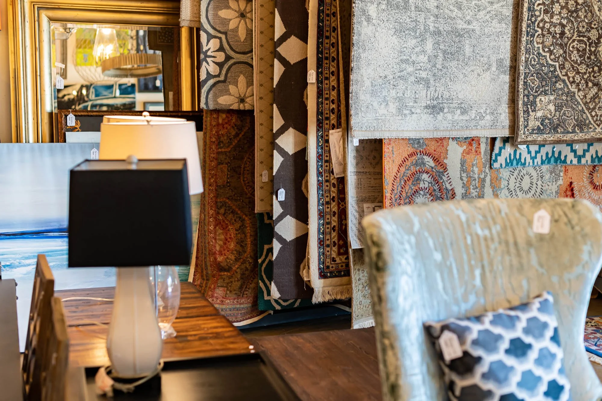Display of various rugs and carpets in a furniture store, with a table and lamps in the foreground.