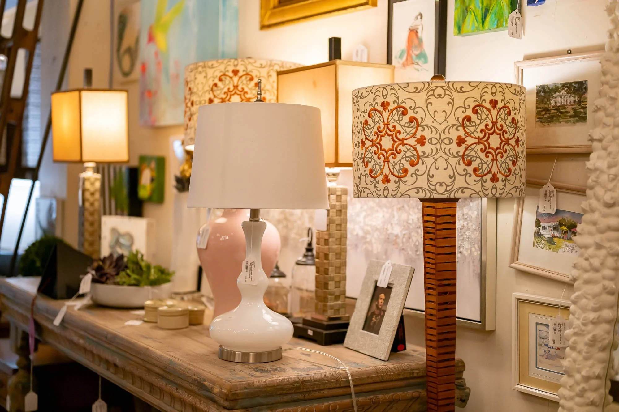 Multiple table lamps of various styles and colors on a wooden surface in an art and decor store, with framed artwork and decorative items in the background.