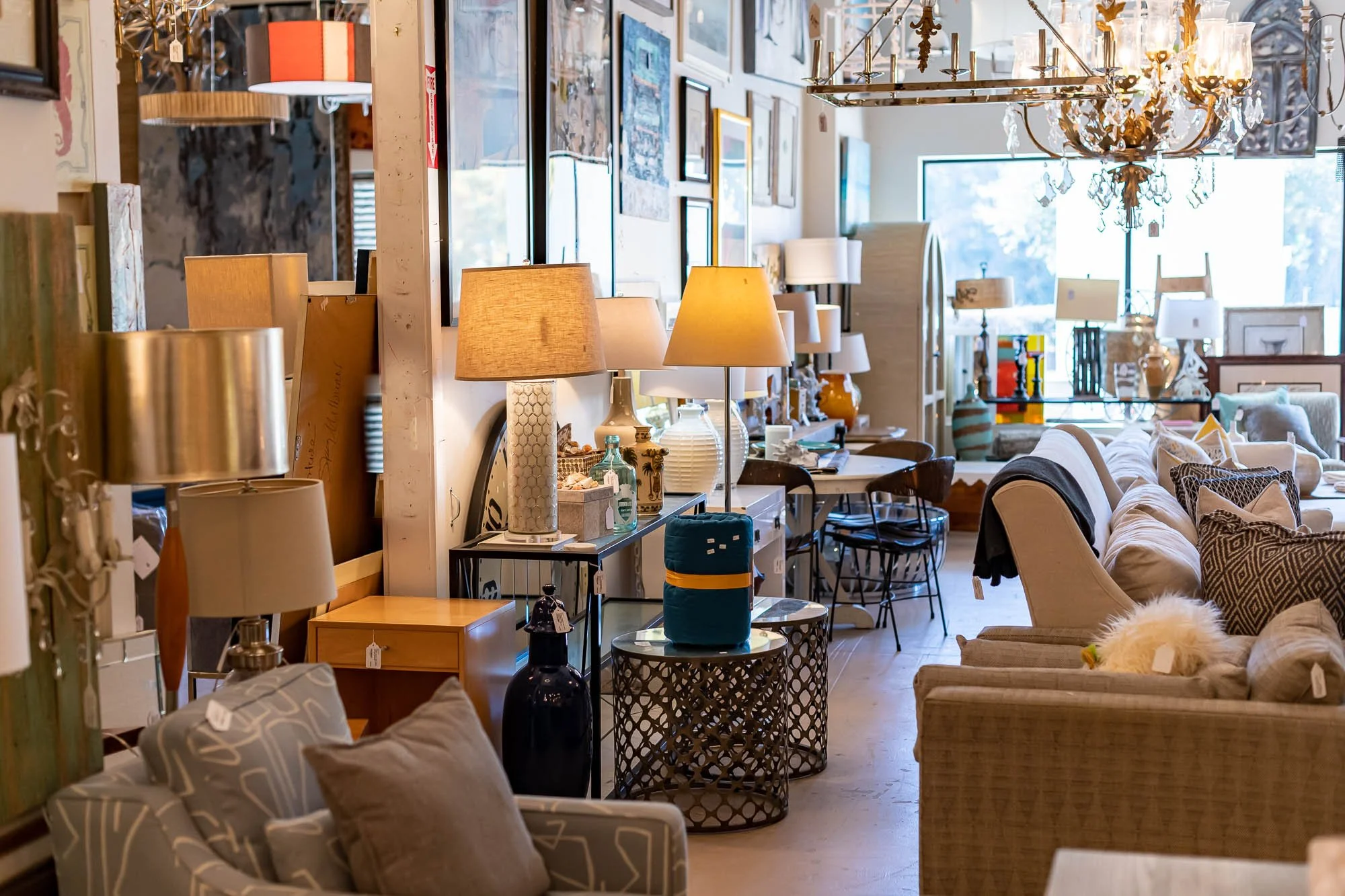 Interior view of a furniture store with sofas, lamps, tables, and decorative items, illuminated by natural light coming through large windows.