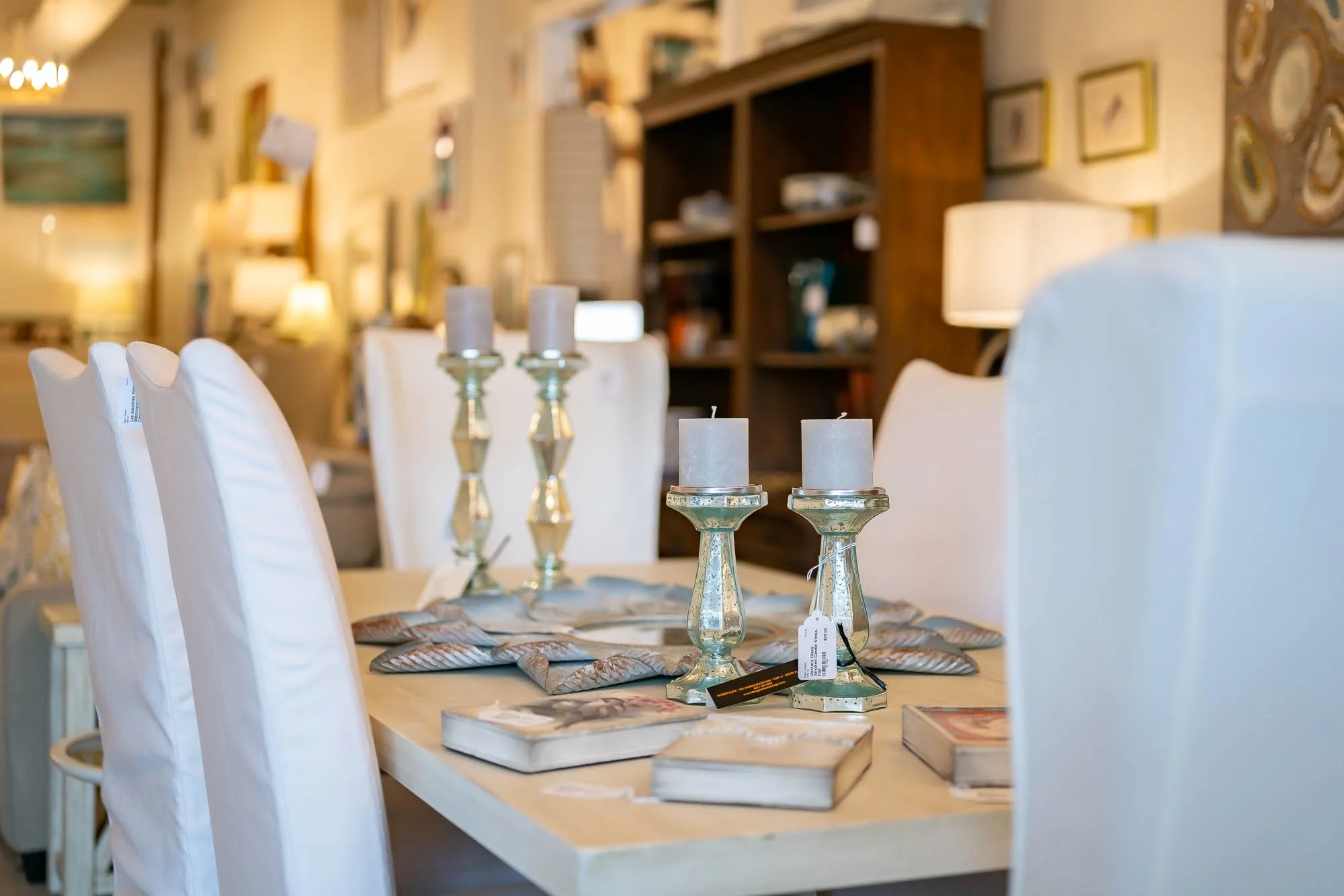 A dining table decorated with three tall candlesticks with candles and books, in a cozy room with framed pictures and lamps.