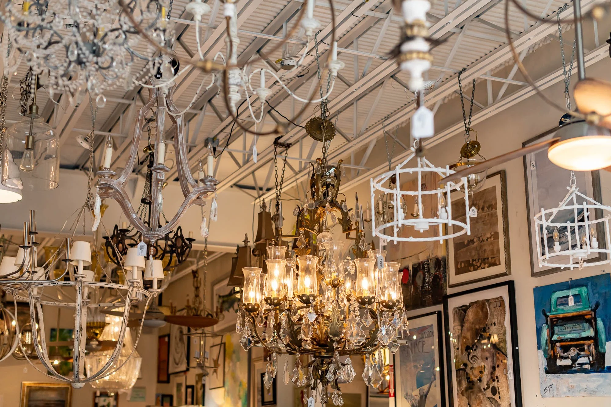 Various ornate chandeliers hanging from a ceiling in a store, with framed artwork on the walls.