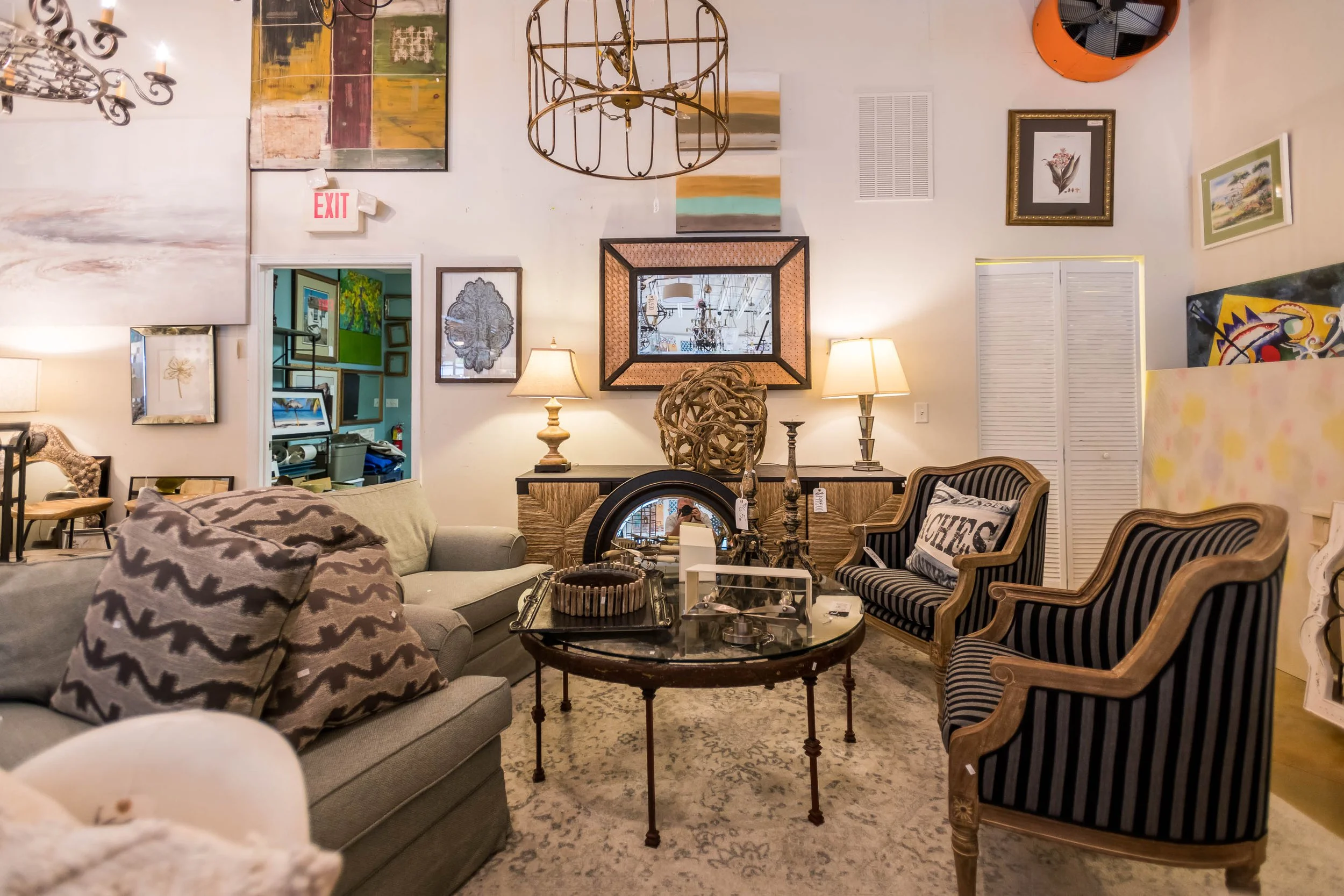Show room with eclectic decor, including framed artwork, lamps, a chandelier, and various furniture pieces like a sofa and striped armchairs.