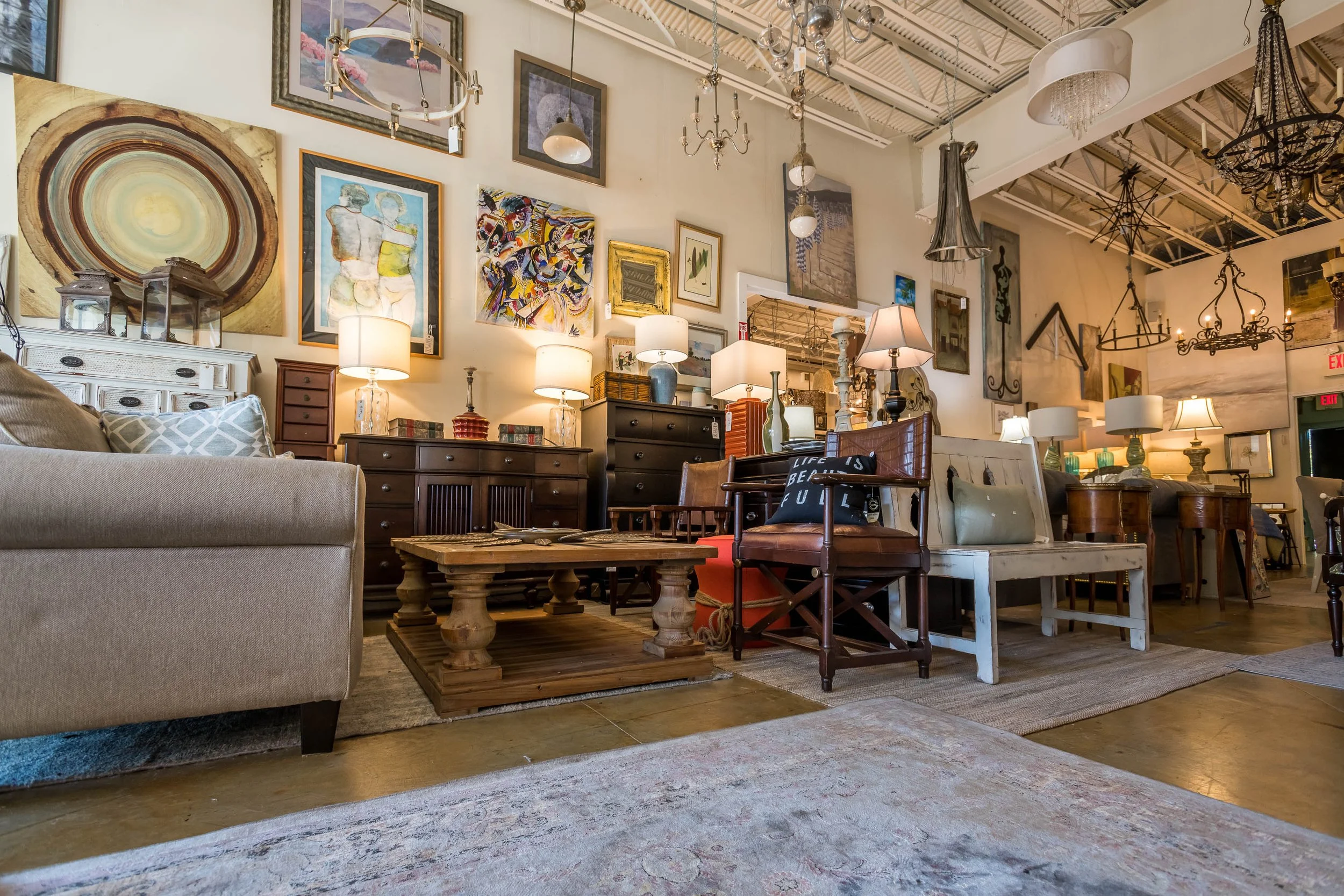 A cozy interior living space with a variety of furniture pieces including sofas, chairs, a wooden coffee table, and plenty of lamps. The wall is decorated with numerous framed artworks and paintings. The ceiling has lighting fixtures and chandeliers,