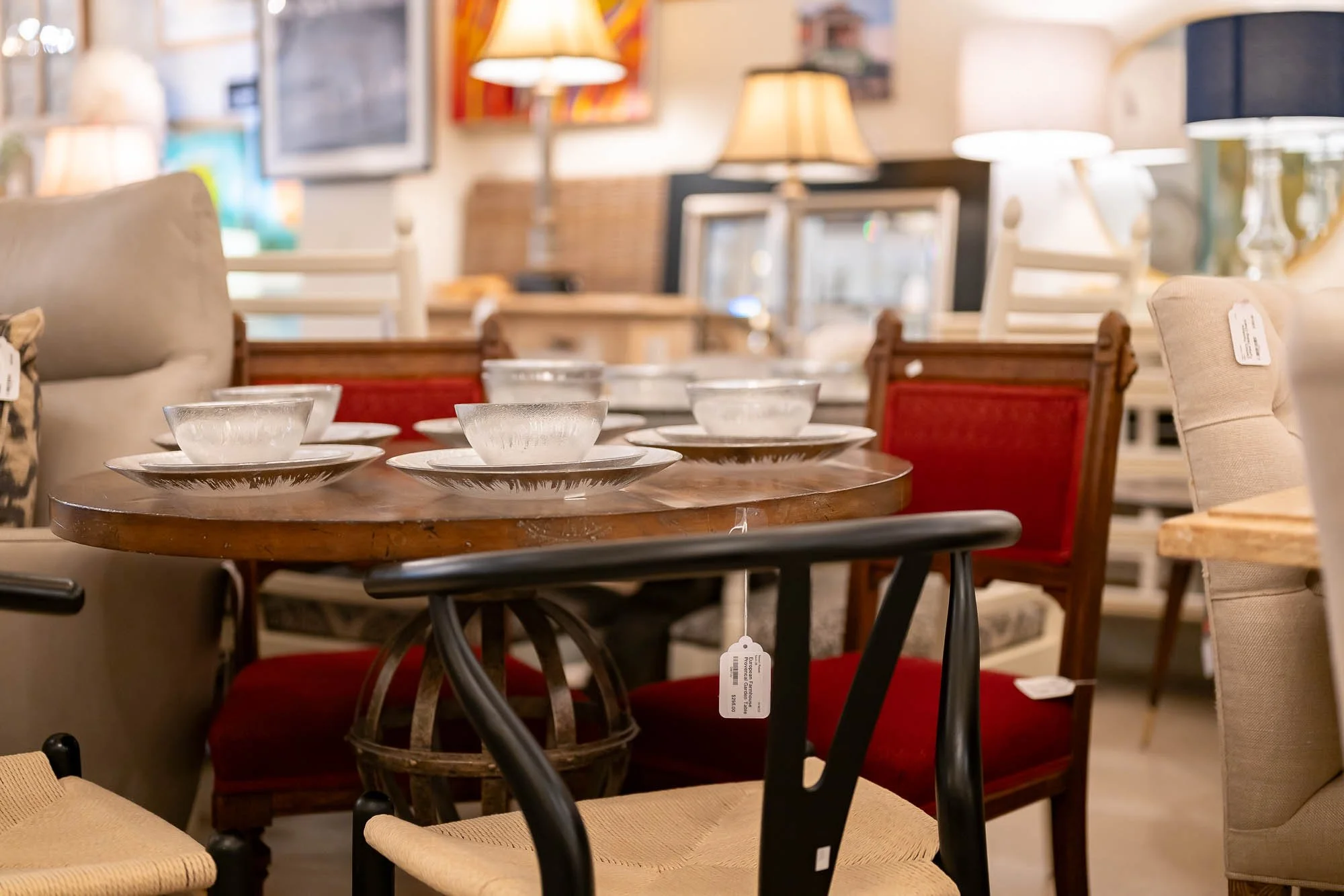 A dining table set with six glass bowls on white plates, surrounded by various chairs in a furniture store with lamps and artwork on the walls.