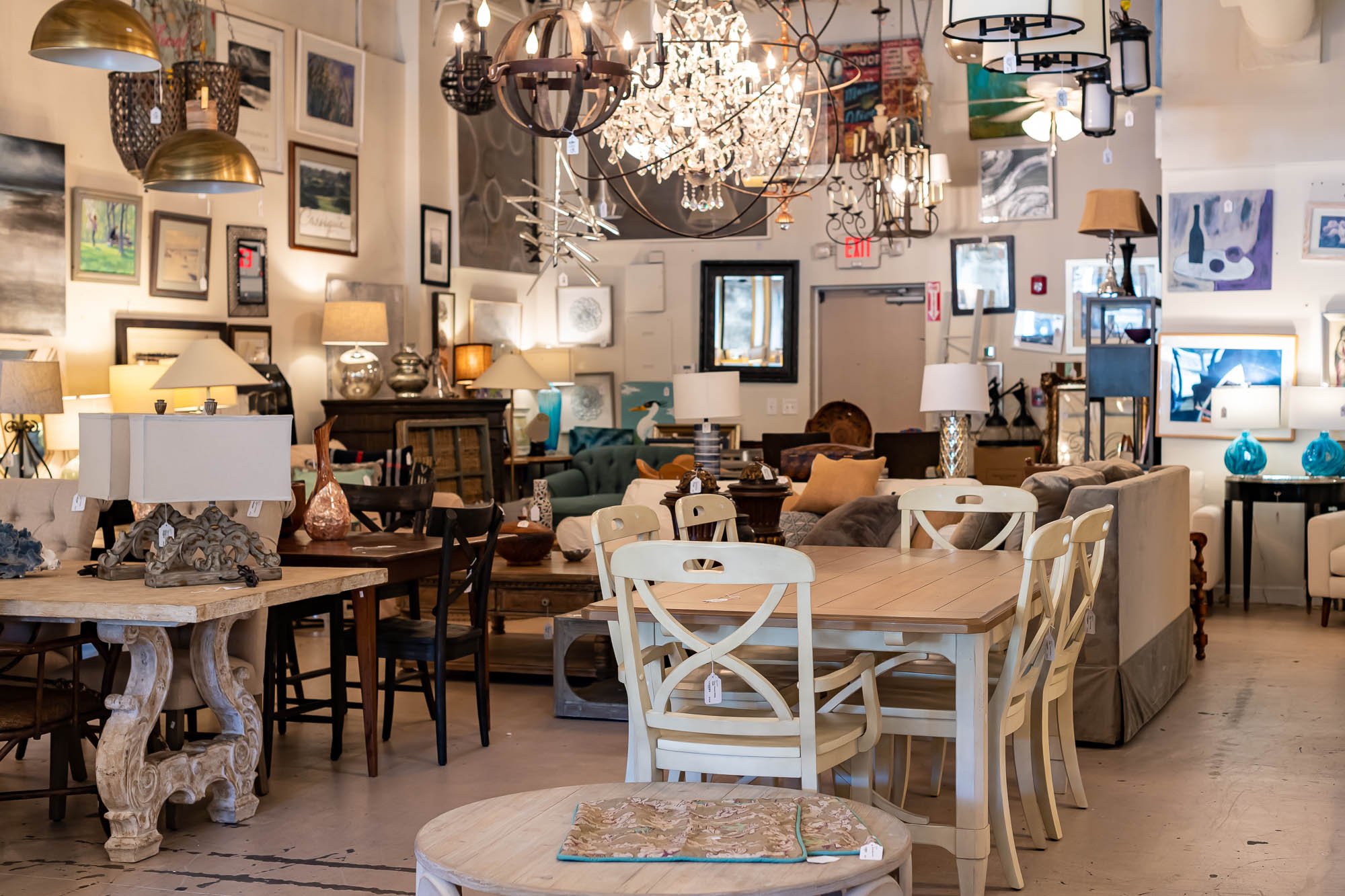 Showroom filled with various furniture and lighting fixtures, including lamps, tables, chairs, and chandeliers, with artwork on the walls.