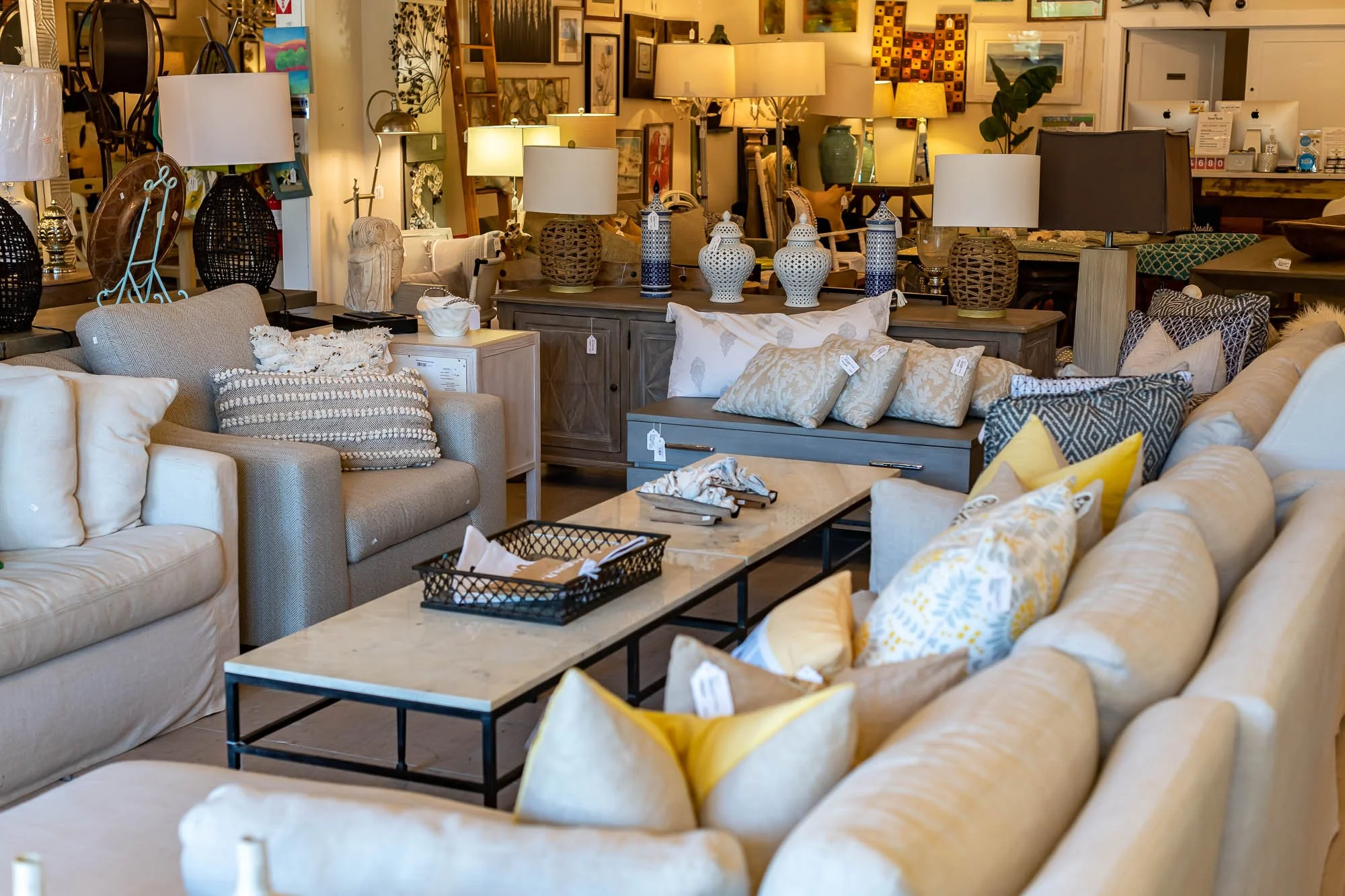 A furniture store displays beige and gray sofas with decorative pillows, a marble-top coffee table, and various lamps and decorative items in the background.