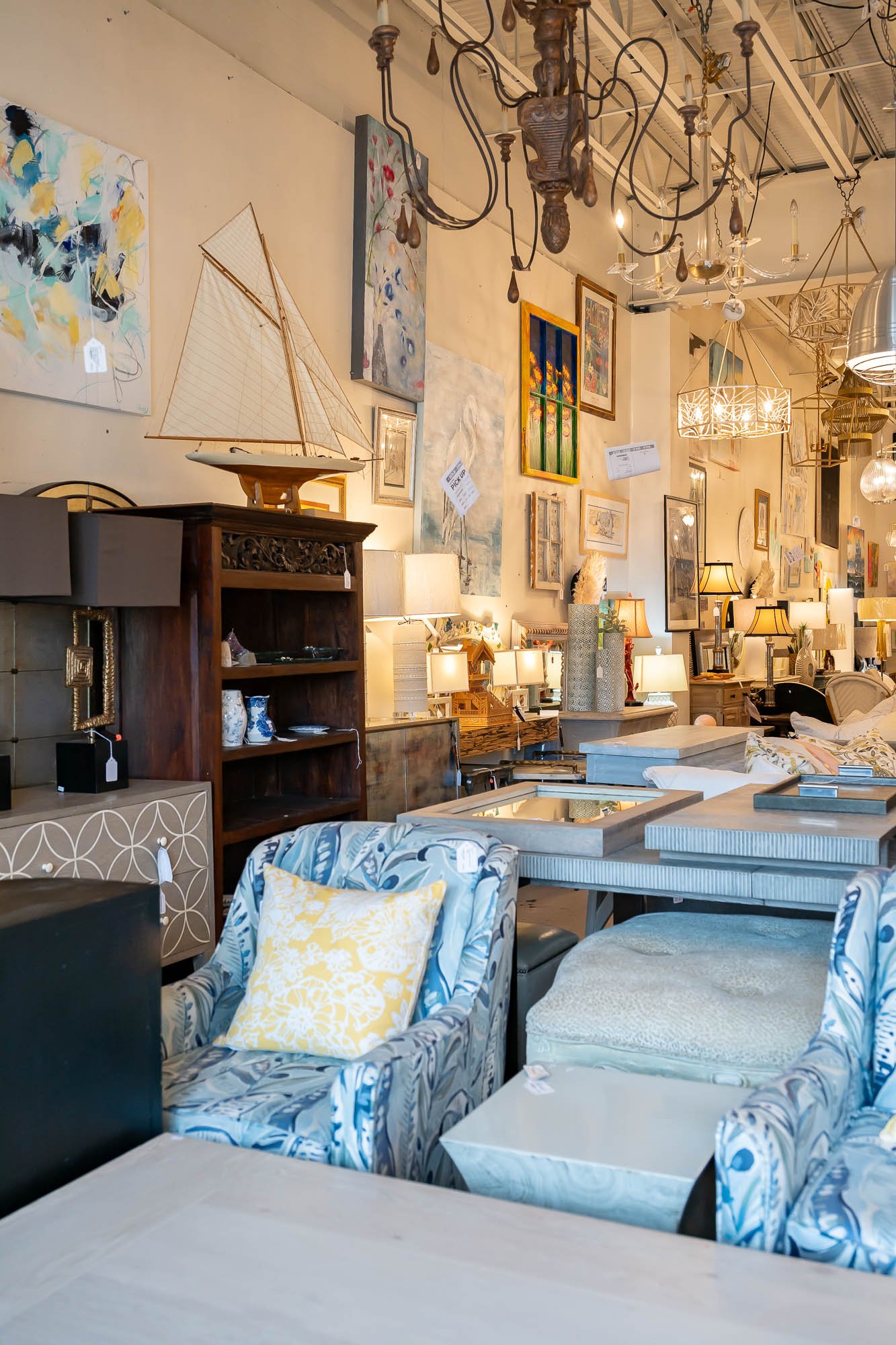 An interior view of a furniture store with various lamps, paintings, and chairs, including an armchair with a yellow pillow and a patterned fabric.