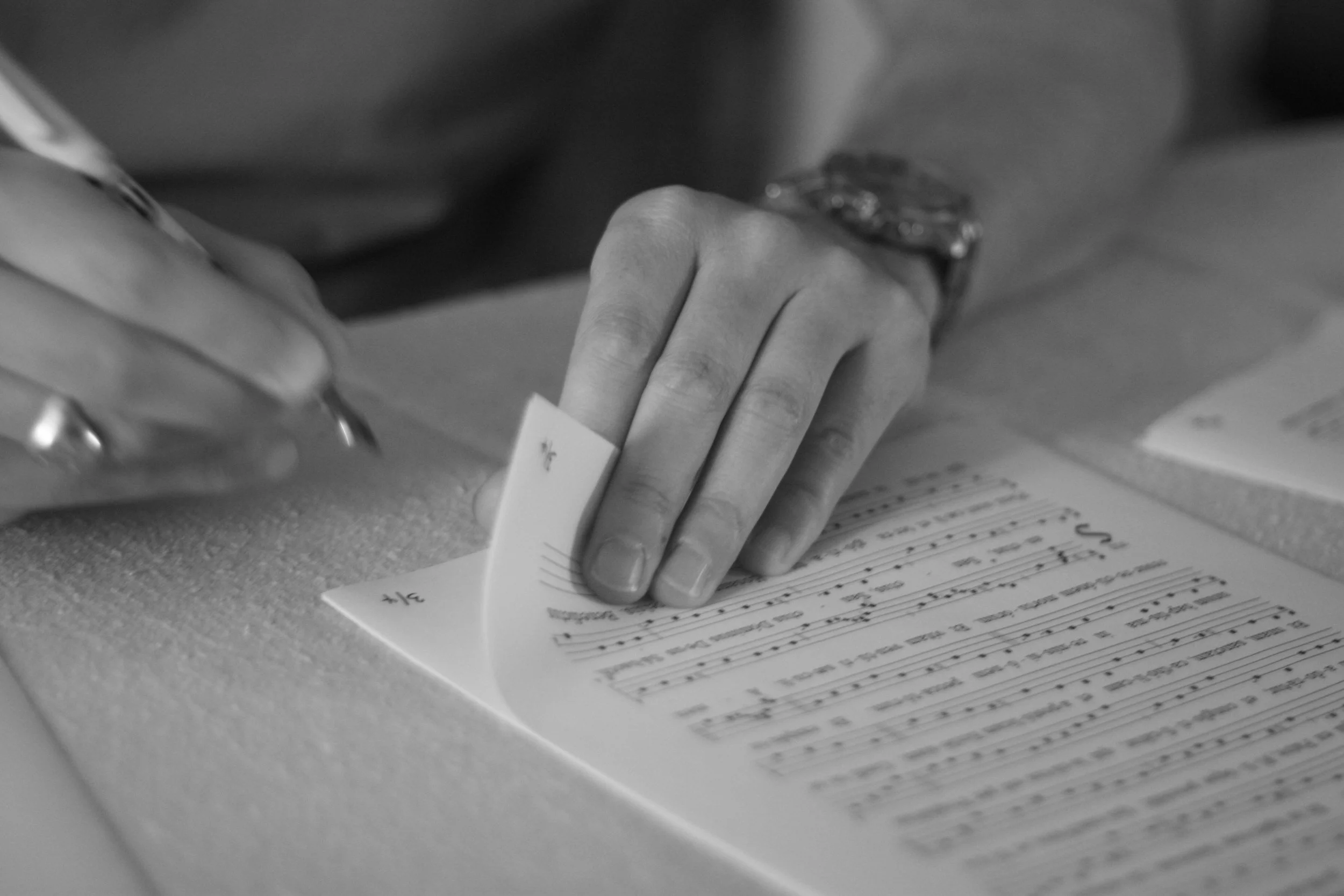 Close-up image of a person's hand with rings on fingers, holding a piece of paper on a desk with handwritten and printed text.