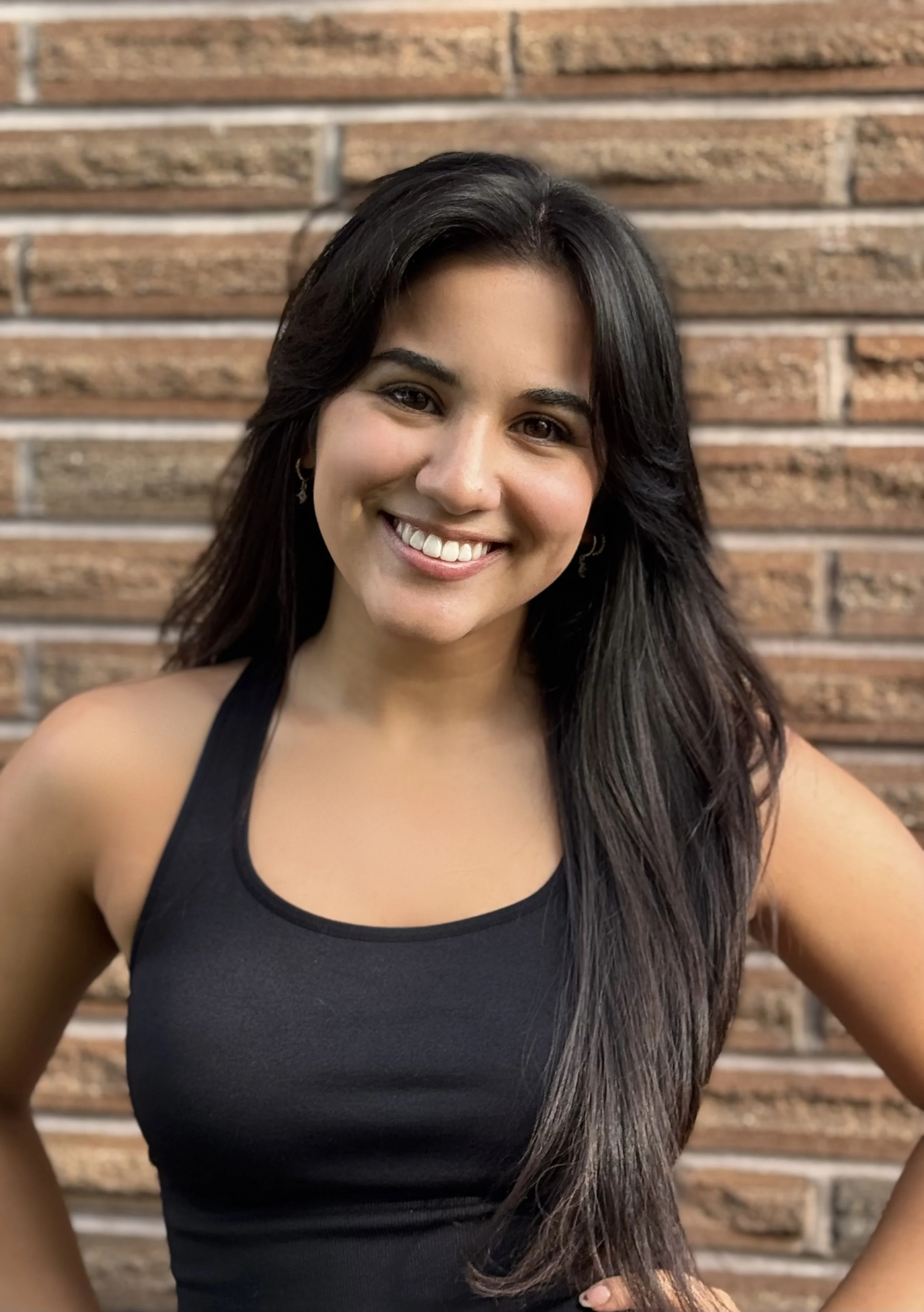 A woman smiling, with long dark hair, wearing a black tank top, standing in front of a brick wall.