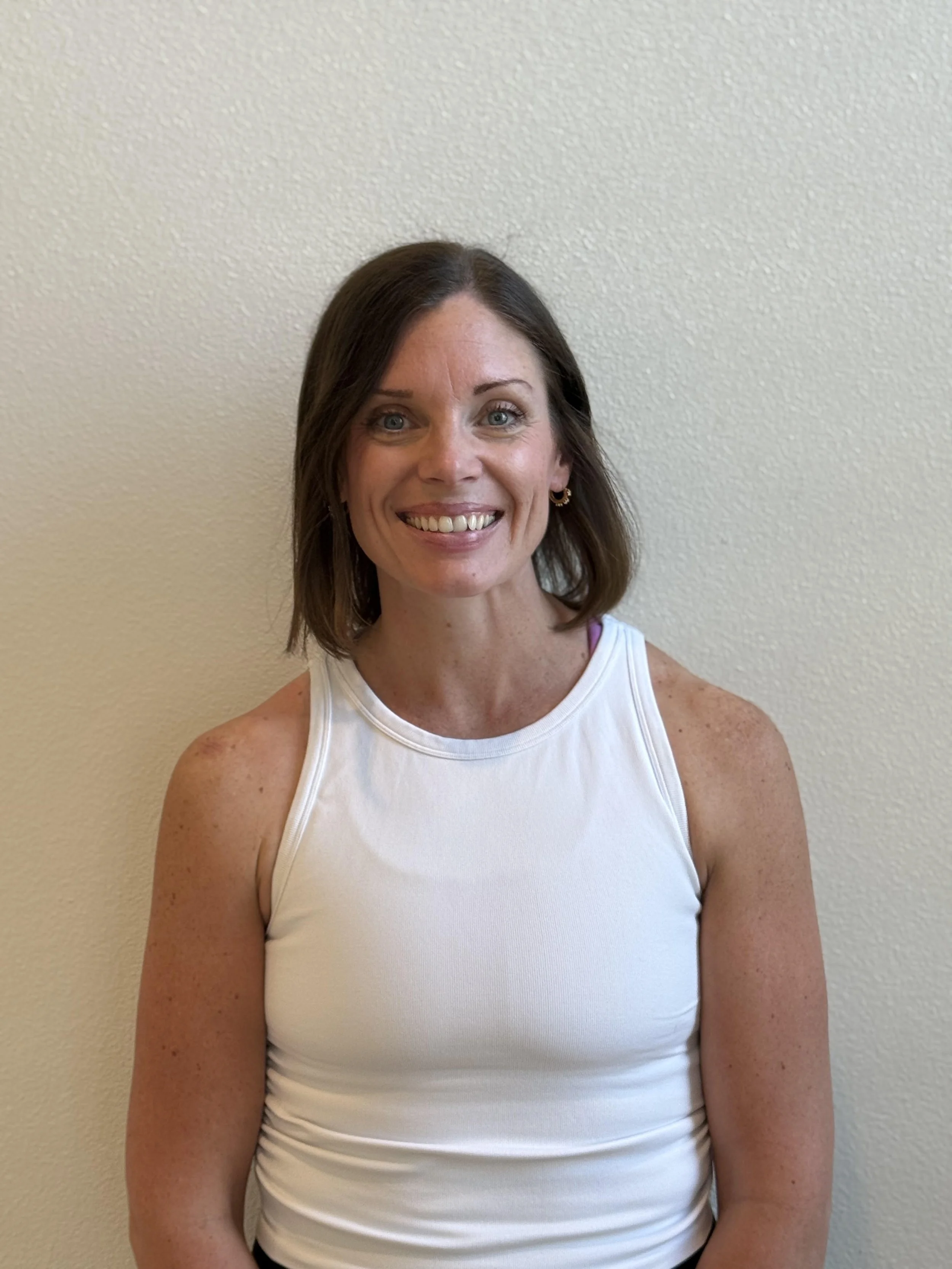 A smiling woman with shoulder-length brown hair and blue eyes, wearing a white sleeveless top, standing against a plain beige wall.